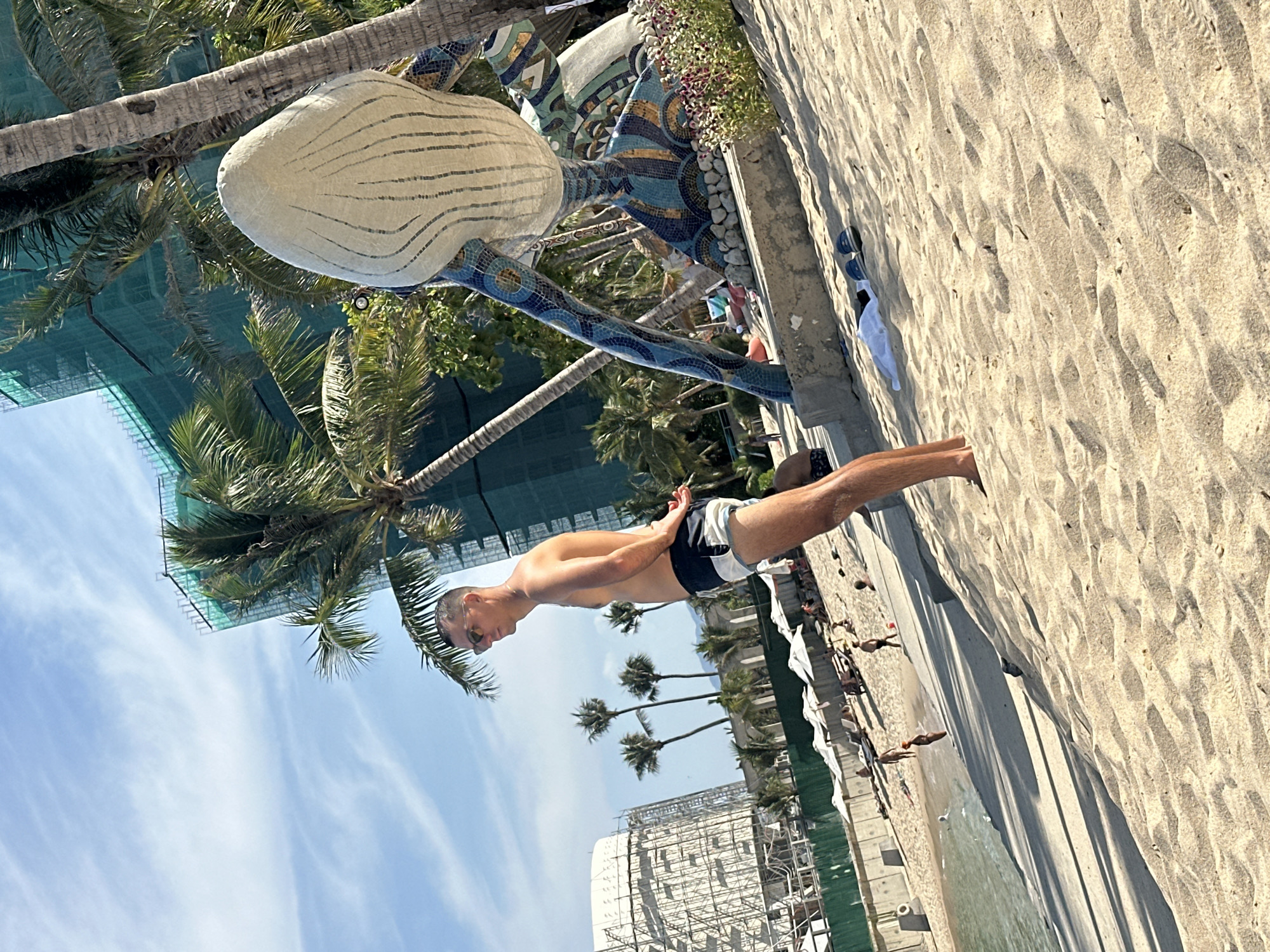 A man is standing on a sandy beach in Bac Nha Trang Ward, Vietnam. He is facing away from the ocean and towards a large, stylized seashell sculpture situated near palm trees. The man is fair-skinned, wearing swimming trunks, and has his hands clasped behind his back. A partly-constructed building can be seen in the background. Other people are scattered along the beach near the water's edge.