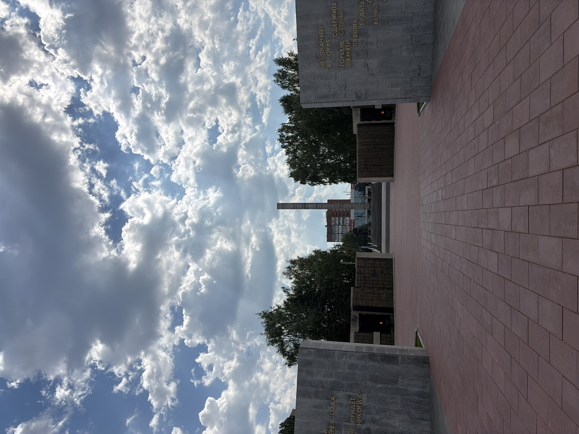 The image depicts a memorial complex, likely dedicated to fallen soldiers, situated in the city of Barnaul, Russia. In the foreground, a red brick paved walkway leads towards a central monument. On either side of the walkway are large, gray stone walls inscribed with text. Between these walls are smaller, dark stone structures, some of which appear to be flame installations, possibly eternal flames. Behind these elements, trees and buildings are visible, with a tall, slender obelisk-like structure in the distance, a common feature in Soviet-era war memorials. The sky above is filled with dramatic, white and gray clouds against a blue backdrop. The overall impression is one of solemnity and remembrance.