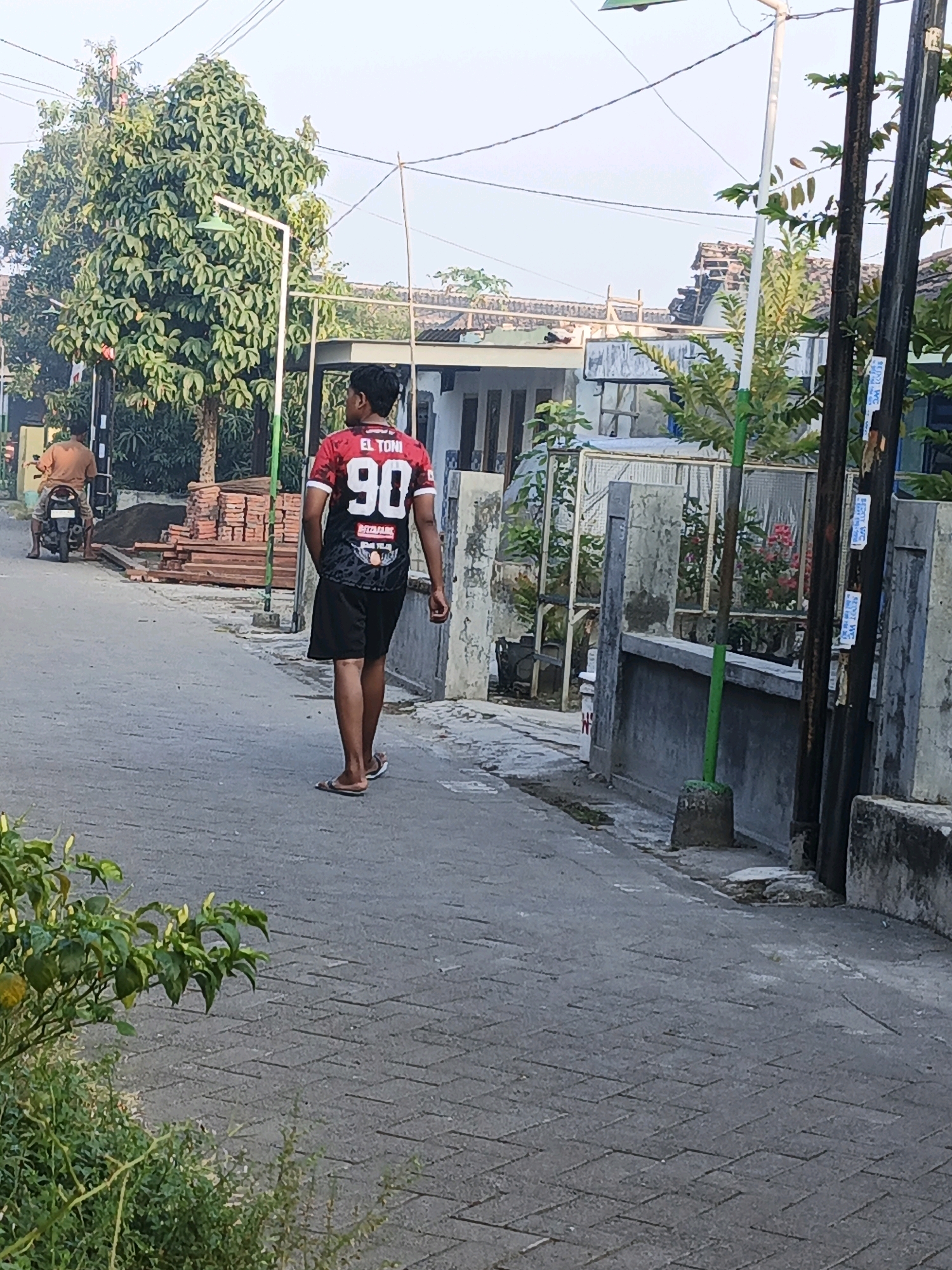 The image depicts a street scene in the city of Ketegan, Indonesia. A young man wearing a red and black jersey with the number "90" and "EL TONI" printed on it, walks away from the camera on a paved street. He is wearing black shorts and sandals. In the background, another person is riding a motorcycle. The street is lined with houses and fences, with trees and vegetation visible. Several utility poles with wires are also present. The overall environment appears to be a residential area.