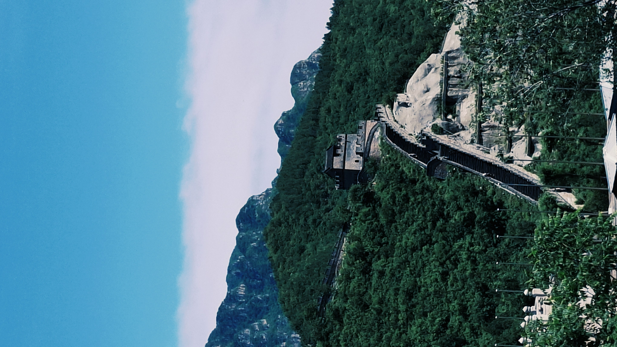 The image shows a portion of the Great Wall of China in the Changping District. The wall snakes along a densely forested mountain ridge. A watchtower is visible, and the wall construction is evident, featuring both stone and brick elements. The sky on the left side is partly clear, with some cloud cover. In the foreground are more trees obscuring the details of the surroundings.