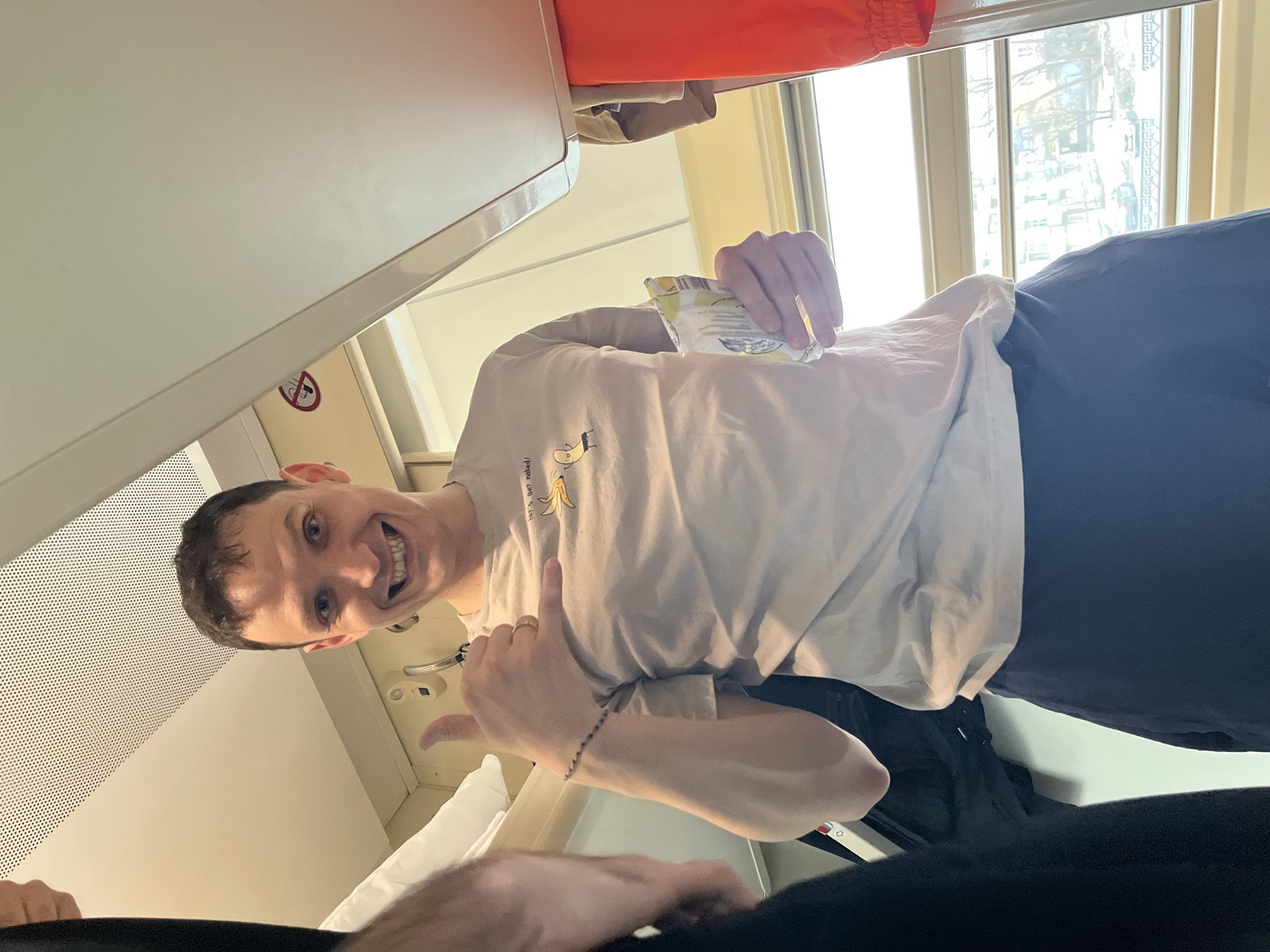 The image shows a smiling man inside what appears to be a train compartment. He's holding a small package in one hand and making a hand gesture with the other. He is wearing a light-colored T-shirt. The compartment has a window to the right, revealing an urban landscape, potentially Izhevsk, Russia. Above the man is the train compartment's overhead storage or ceiling. Below him, other passengers are visible. A pillow is also visible behind his hand.