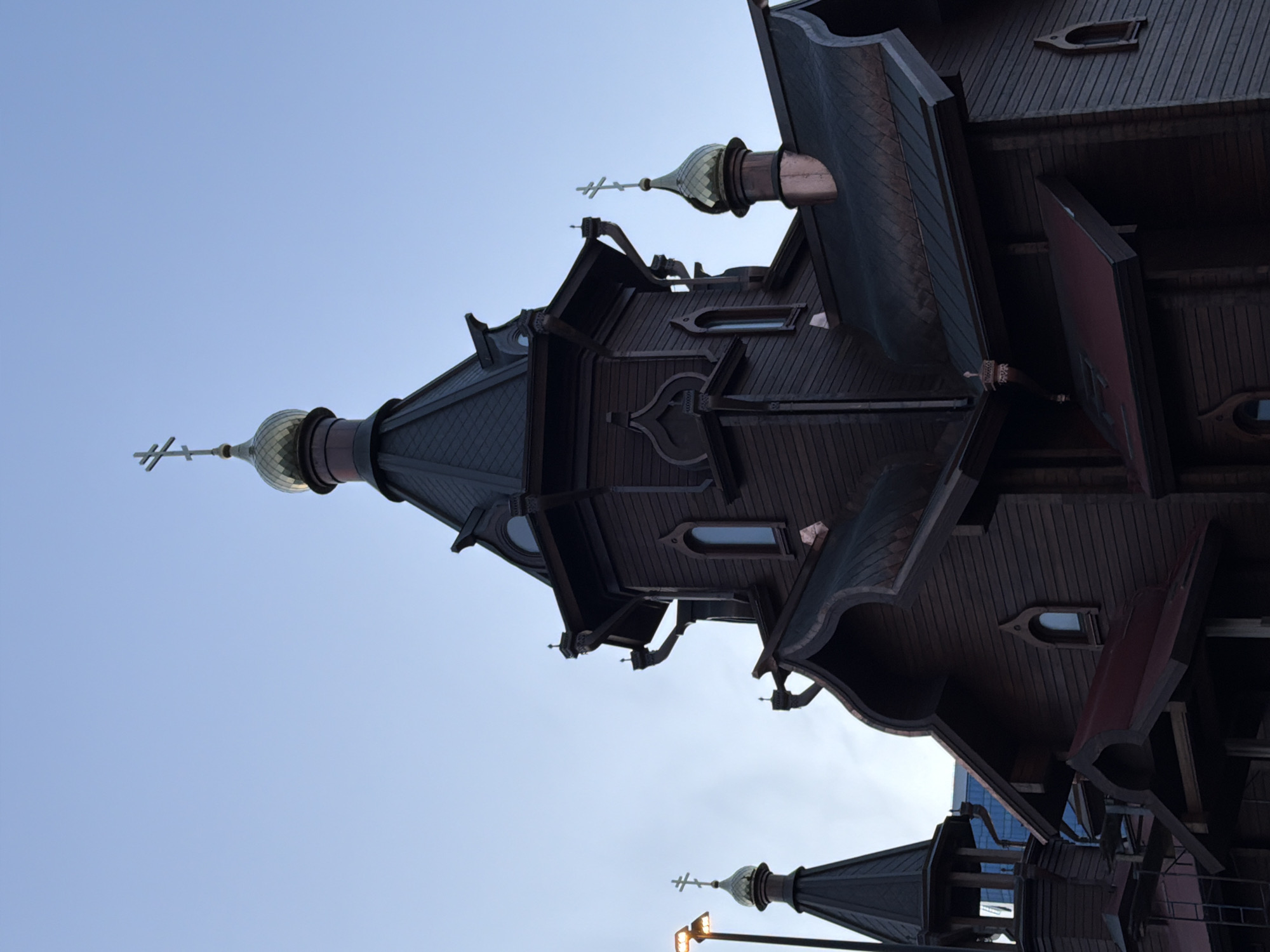 The image displays a large, intricately designed wooden building, likely a church or chapel, against a clear, bright blue sky. The original image is rotated 90 degrees counter-clockwise.
The primary subjects are the architectural elements of the building, which exhibits a distinctive traditional Russian style. Multiple levels and complex rooflines are visible, crafted from dark, horizontally paneled wood. Two prominent onion-shaped domes, each topped with an Orthodox cross, are featured. The larger dome on the left half of the image is metallic-toned, possibly green or bronze, and sits atop a dark spire. A smaller dome and cross are partially visible towards the bottom right.
Ornate details, including carved patterns, decorative brackets, and unique window shapes (some arched, some rectangular), adorn the wooden facade. Some elements, particularly on the roofing and decorative accents, appear to have a copper finish. The building is viewed from below, looking upwards, emphasizing its height and detail against the unblemished sky.
No people are visible, and no specific activity is taking place. The clear blue sky indicates bright daytime conditions, likely morning or afternoon. The architectural style and the presence of Orthodox crosses are consistent with the provided location context of Krasnoyarsk, Russia. There is no visible text in the image.