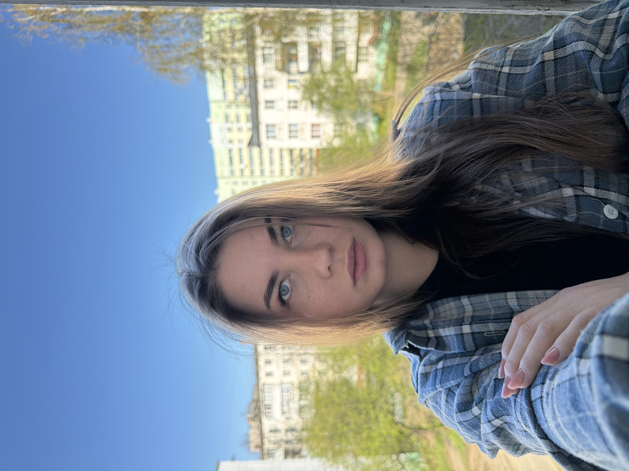 A young woman poses for a selfie against an outdoor backdrop. She has light skin, blue eyes, and long brown hair with blonde highlights near her face. She wears a black shirt under a blue and white plaid flannel shirt. Her hand, adorned with pink nails, is visible in the lower right corner.
In the blurred background are green trees, a clear blue sky, and a multi-story apartment building. This location is likely Chaikovsky, Russia, given the prompt. The focus is on the woman's face and upper body. The photograph is oriented vertically.