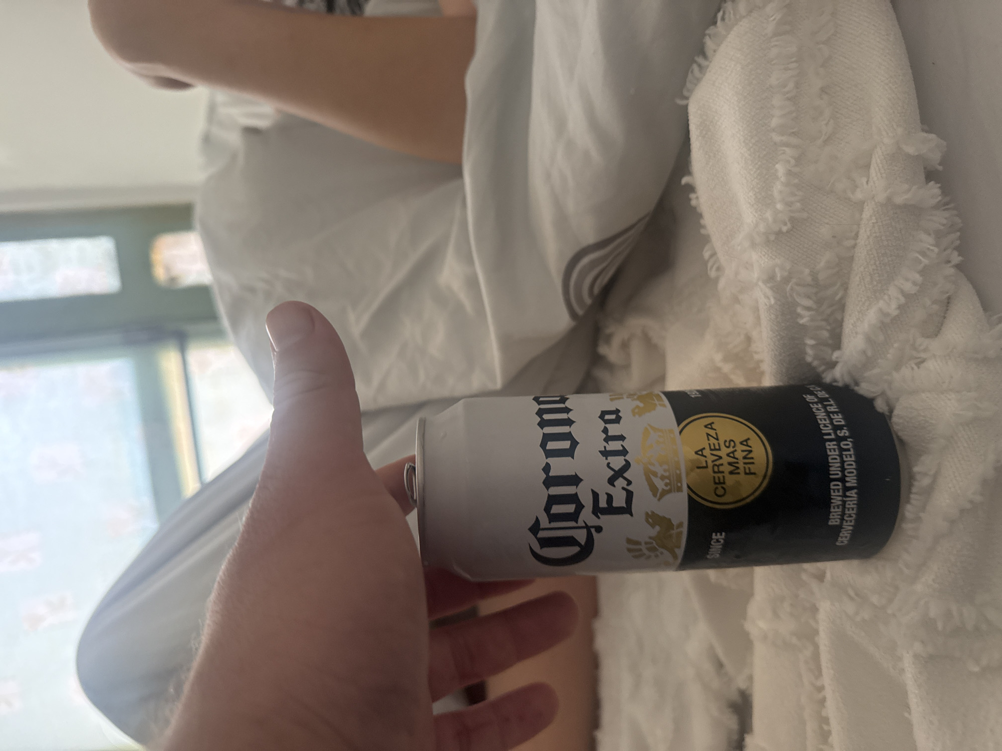 In Herzliya, Israel, a person's hand is positioned over a can of Corona Extra beer. The can is silver with a black bottom and features the brand's logo and crown imagery. The person's hand, with visible fingernails and slight hair on the arm, is resting on the top of the can, with a thumb and index finger lightly touching it. The background suggests an indoor setting, possibly a bedroom, with a person lying down covered in white bedding. The overall environment appears relaxed and casual.