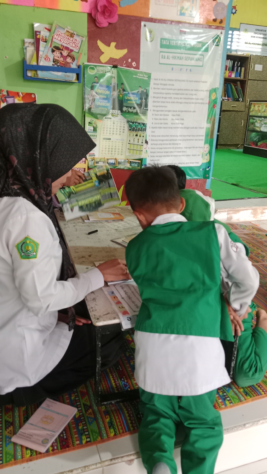 The image depicts a classroom setting in RA Al-Hikmah Sepanjang, Karangharjo, Indonesia. Three individuals are present: an adult woman, likely a teacher, wearing a hijab and a white uniform with a green emblem, and two young boys in green uniforms. The teacher is seated at a low table, holding a book open and appearing to interact with one of the boys, who is also looking at the book. The second boy is facing away from the camera, seated on the floor. The environment includes educational materials on the wall, such as posters and a calendar, and a colorful rug on the floor. The overall scene suggests an educational activity or lesson taking place.