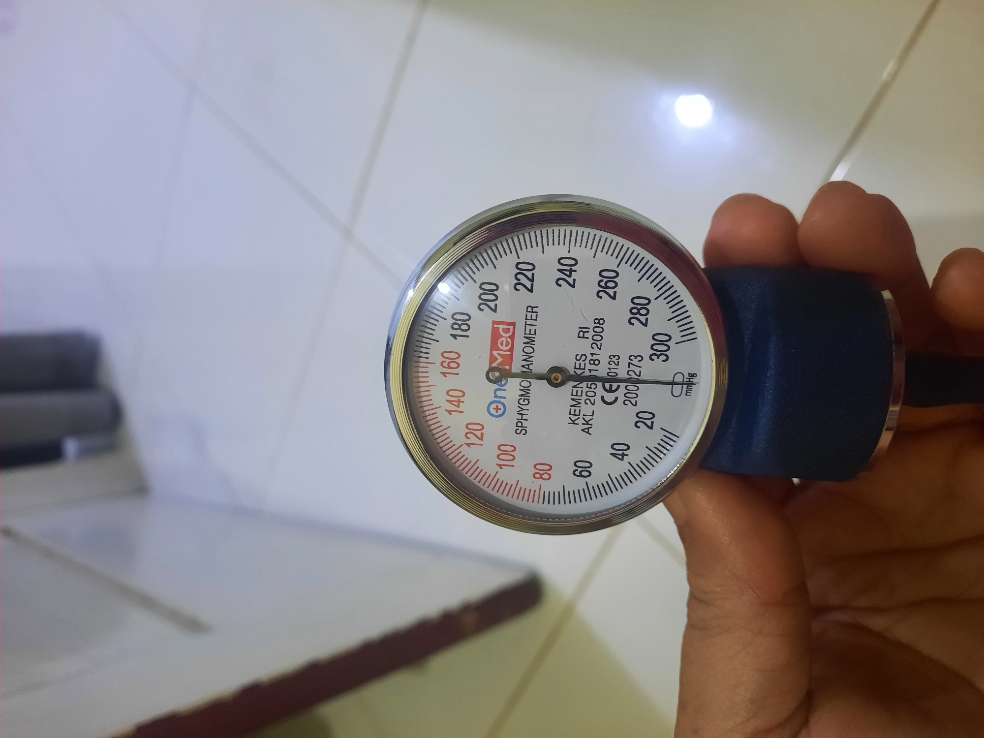 A close-up, top-down, medium shot shows a person's hand holding a sphygmomanometer. The sphygmomanometer is a medical device used to measure blood pressure, with a circular dial marked with numbers from 20 to 300 in increments of 20, and smaller increments of 10. The dial features the brand name "OneMed" in blue and red text. The device also displays regulatory markings, including "KEMENKES RI", "AKL 2051812008", "CE 0123", and "200 273". The measurement unit "mm Hg" is also visible. The background consists of a light-colored tiled floor and a partially visible white wall, suggesting an indoor environment. The lighting is bright, with a noticeable glare on the wall. The scene is devoid of any identifiable landmarks or features that would indicate a specific location within Lhokseumawe, Indonesia, or any city for that matter.