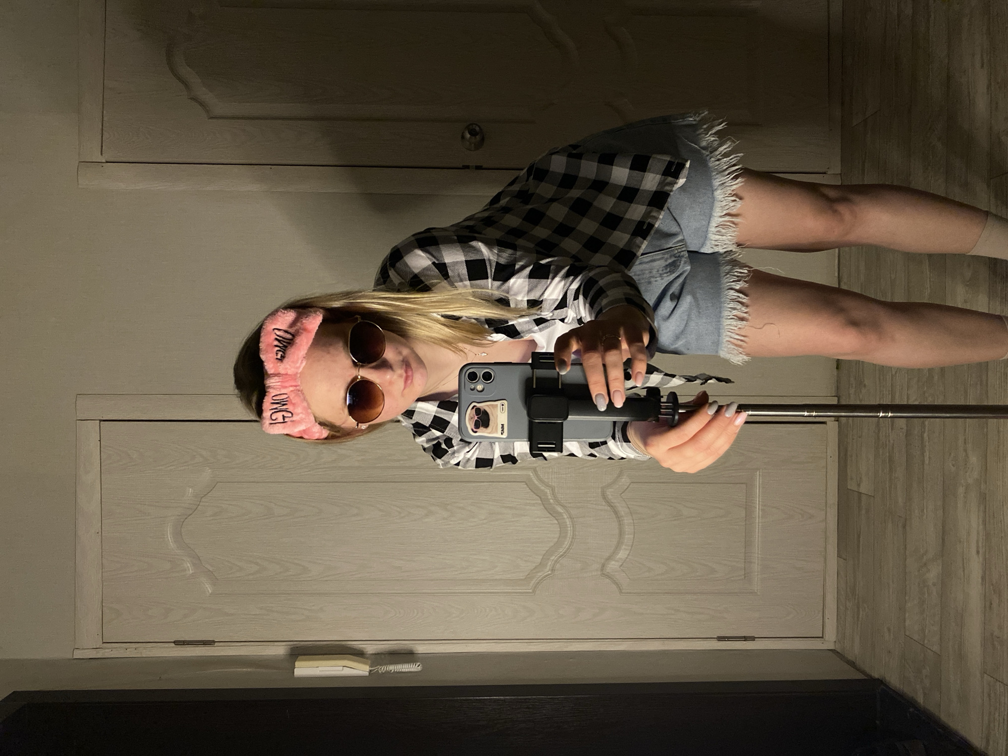 An individual is depicted indoors, taking a selfie in the city of Unknown, Unknown. A light-skinned female with blonde hair, obscured by dark sunglasses, leans forward while holding a smartphone attached to a black selfie stick or tripod. She wears a pink terry cloth headband with "OMG" written in reverse, a black and white plaid button-up shirt over a light-colored top, light blue frayed denim cutoff shorts, and light-colored socks. Her fingernails are painted light grey, and she wears a necklace and a ring on her right hand. The smartphone, an iPhone with a dark grey case, features a pop socket or grip with a cartoon pug wearing sunglasses and "SNM" text. The background consists of light grey/beige walls and light wood-grain flooring. Two light-colored doors with decorative raised panels are visible; the left door shows a hinge, and the right door has a round doorknob. A white wall-mounted intercom or telephone unit with a coiled cord is present on the left wall. The scene is dimly lit with artificial light.