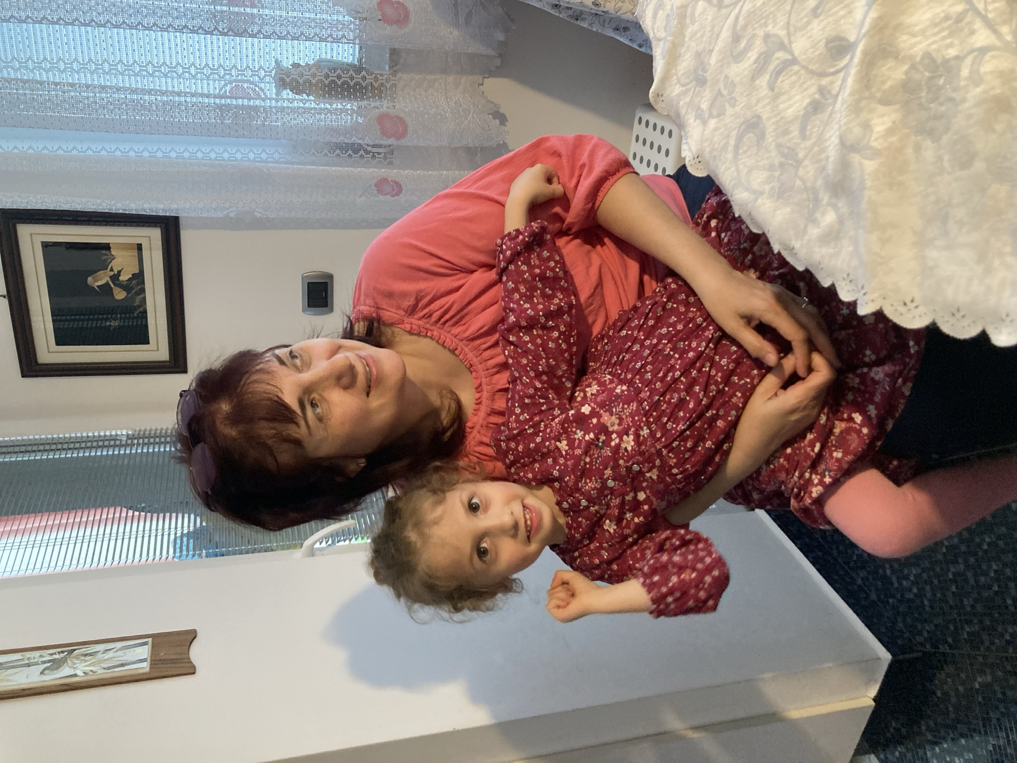 In a residential interior, a woman with shoulder-length reddish-brown hair, wearing a pink blouse, holds a young girl with curly blonde hair. The girl is dressed in a burgundy floral-patterned dress and pink leggings. Both are smiling and looking towards the camera. The woman's left hand rests on the girl. A white lace-edged tablecloth is partially visible in the upper right. Background details include a framed picture on a wall and a window with white curtains. The city and specific location are unknown.