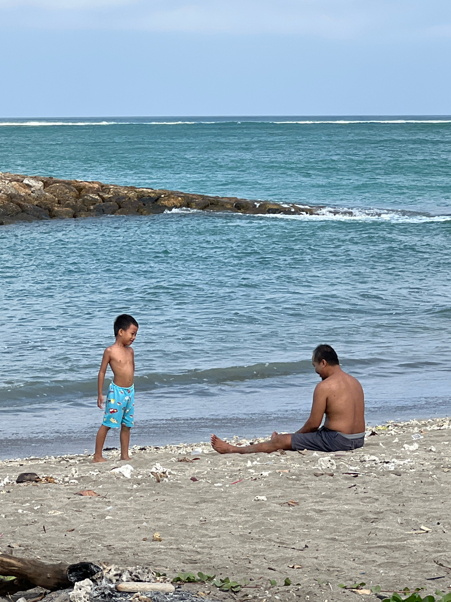 A boy and a man are on the beach in Kuta, Indonesia. The boy is standing, facing the ocean, wearing blue swim trunks with a pattern of cars. The man is sitting on the sand, facing away from the camera, wearing gray shorts. The ocean is calm with small waves, and there is a stone jetty in the background. The beach is sandy and scattered with debris, including shells and driftwood. The sky is overcast.