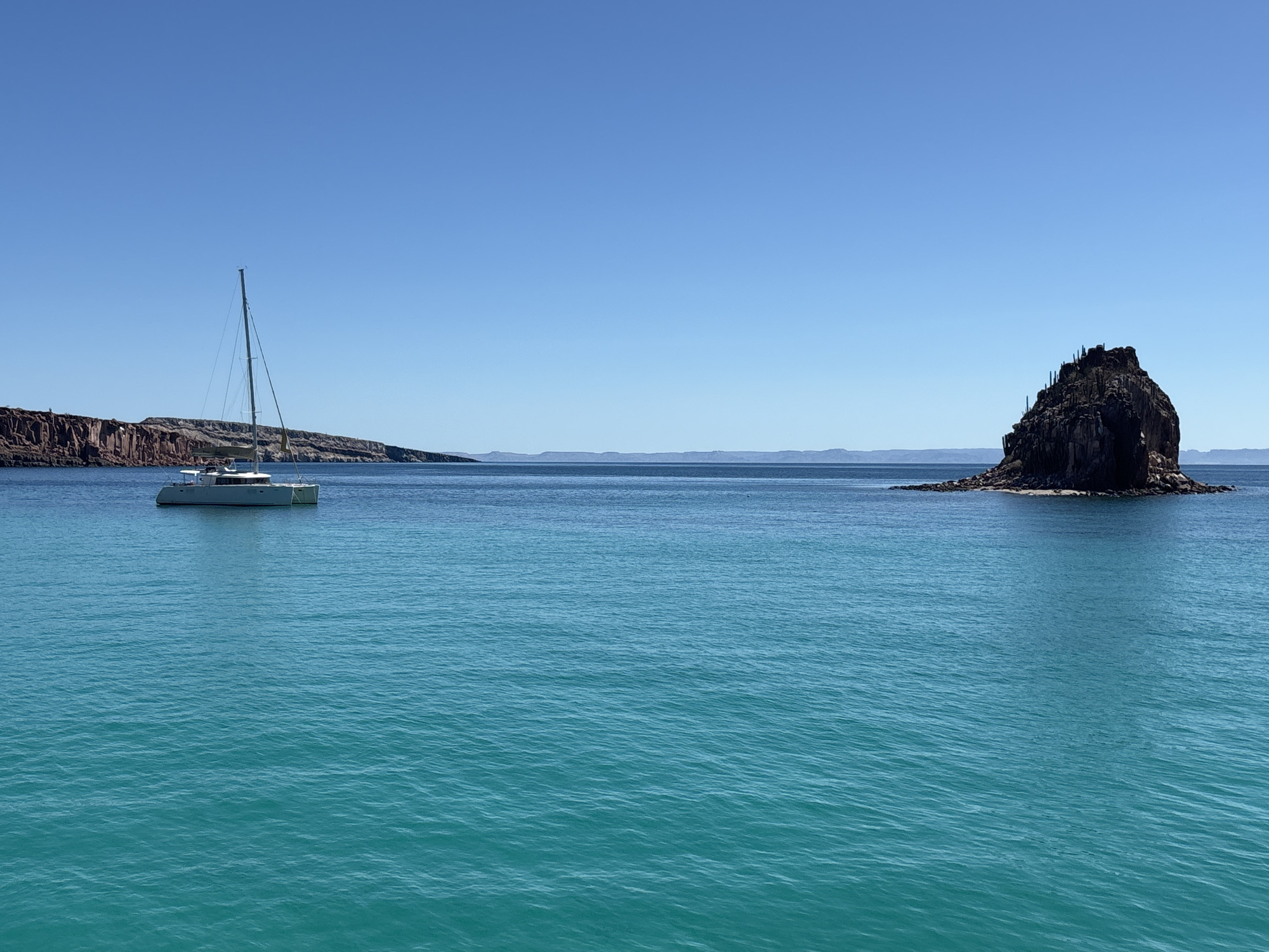 The image presents a coastal scene in what is described to be the city of Unknown, Mexico. The scene features a clear blue sky above a tranquil body of water, a turquoise sea that extends to the horizon. A white catamaran sailboat is positioned on the left side of the frame, anchored or drifting near the shoreline. A rocky islet, capped with sparse vegetation or cacti, stands prominently to the right. In the background, a mountainous coastline can be discerned. The overall impression is one of serenity and natural beauty.