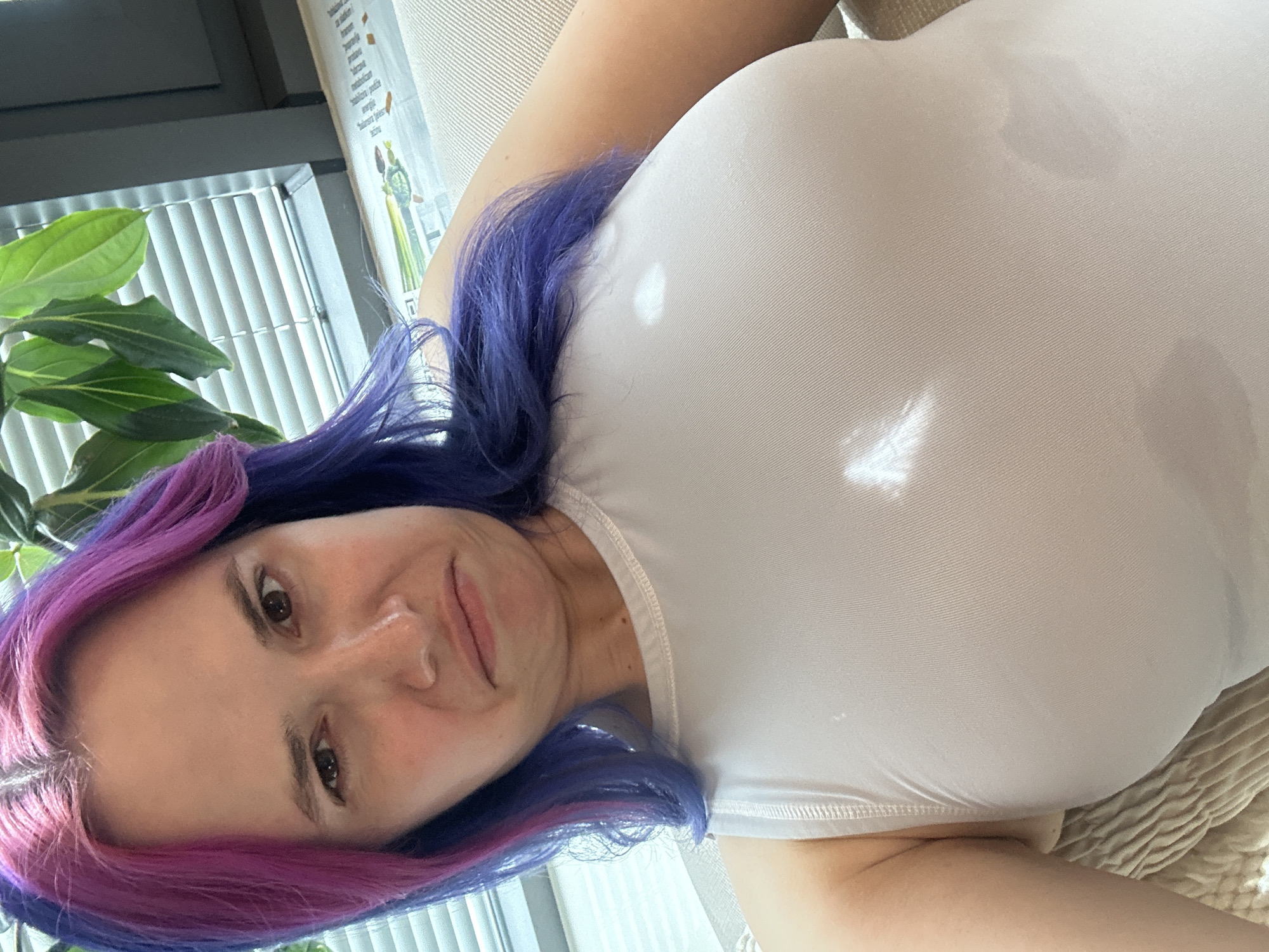A woman with striking, two-toned hair, vibrant purple transitioning to pink, is captured in a close-up indoor shot. She is lying on her side, looking directly at the camera with a gentle, friendly smile. She wears a plain white, form-fitting top, which exhibits natural light reflections from an external source.
The setting is an interior space, likely a room with a window that lets in ample daylight. Through the window, vertical blinds are visible. To the left of the woman, a lush green houseplant with large leaves is partially in view. Behind her, on the wall, there is a poster or chart. Though partially obscured, several Croatian words related to health and nutrition can be discerned, such as "zdravlje" (health), "hrana" (food), "voće" (fruit), "povrće" (vegetables), "minerali" (minerals), "vitamini" (vitamins), "voda" (water), and "energija" (energy). This detail aligns with the provided location context of the City of Zagreb, Croatia.
A textured, light-colored cushion or blanket is visible beneath her, suggesting a comfortable, relaxed position. The lighting indicates it is daytime. The image conveys a casual and personal moment.