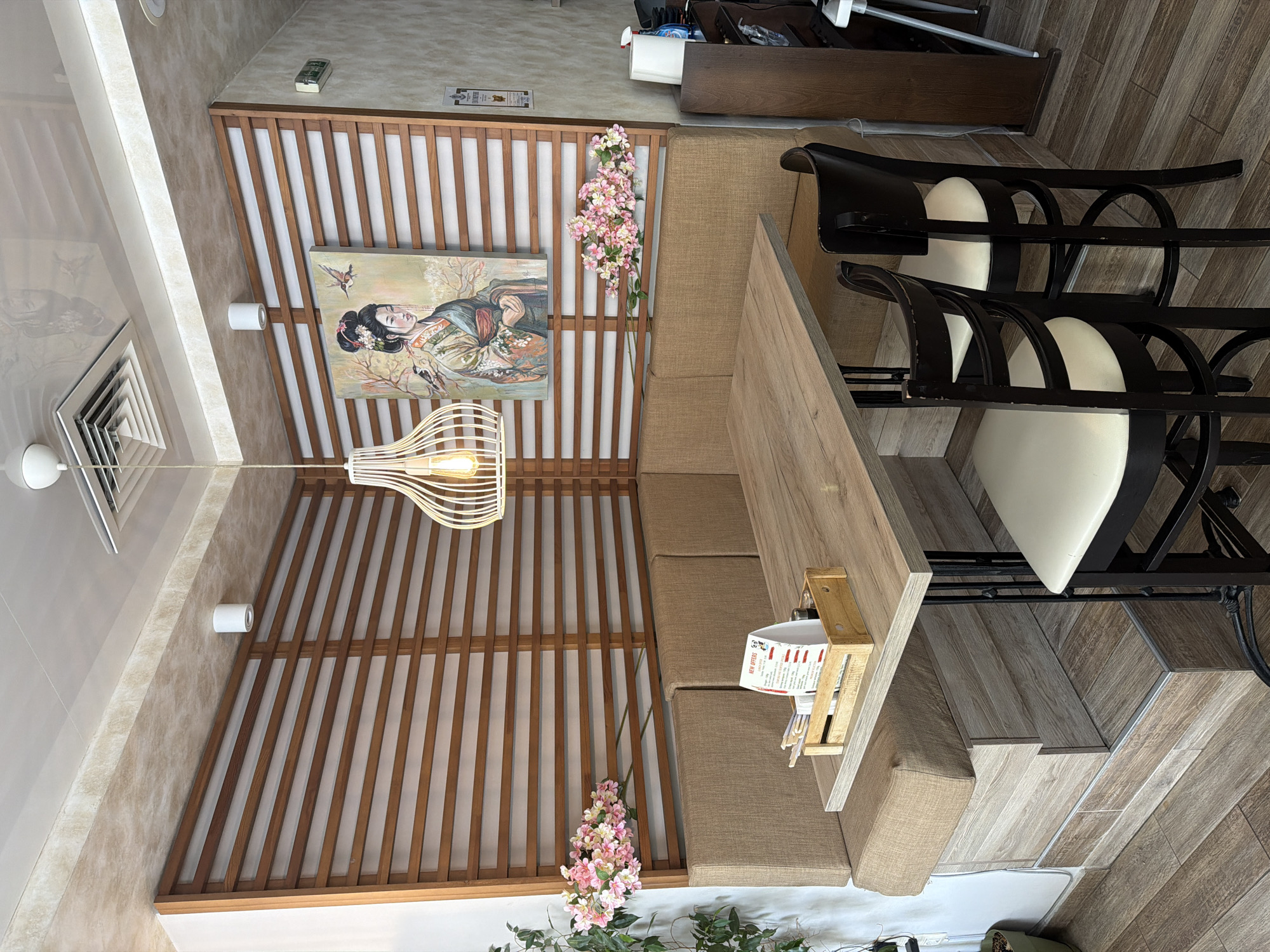 The image shows the interior of a restaurant, possibly located in Varna, Bulgaria. The scene is a dining area with a table and chairs. The table is set against a booth-style seating arrangement. The chairs are black with white seats and are positioned on a wooden-tiled floor. The ceiling has a decorative wooden slat design with an illustration of a geisha. Floral decorations are present near the ceiling, and a modern light fixture hangs above the table. A menu holder is visible on the table.