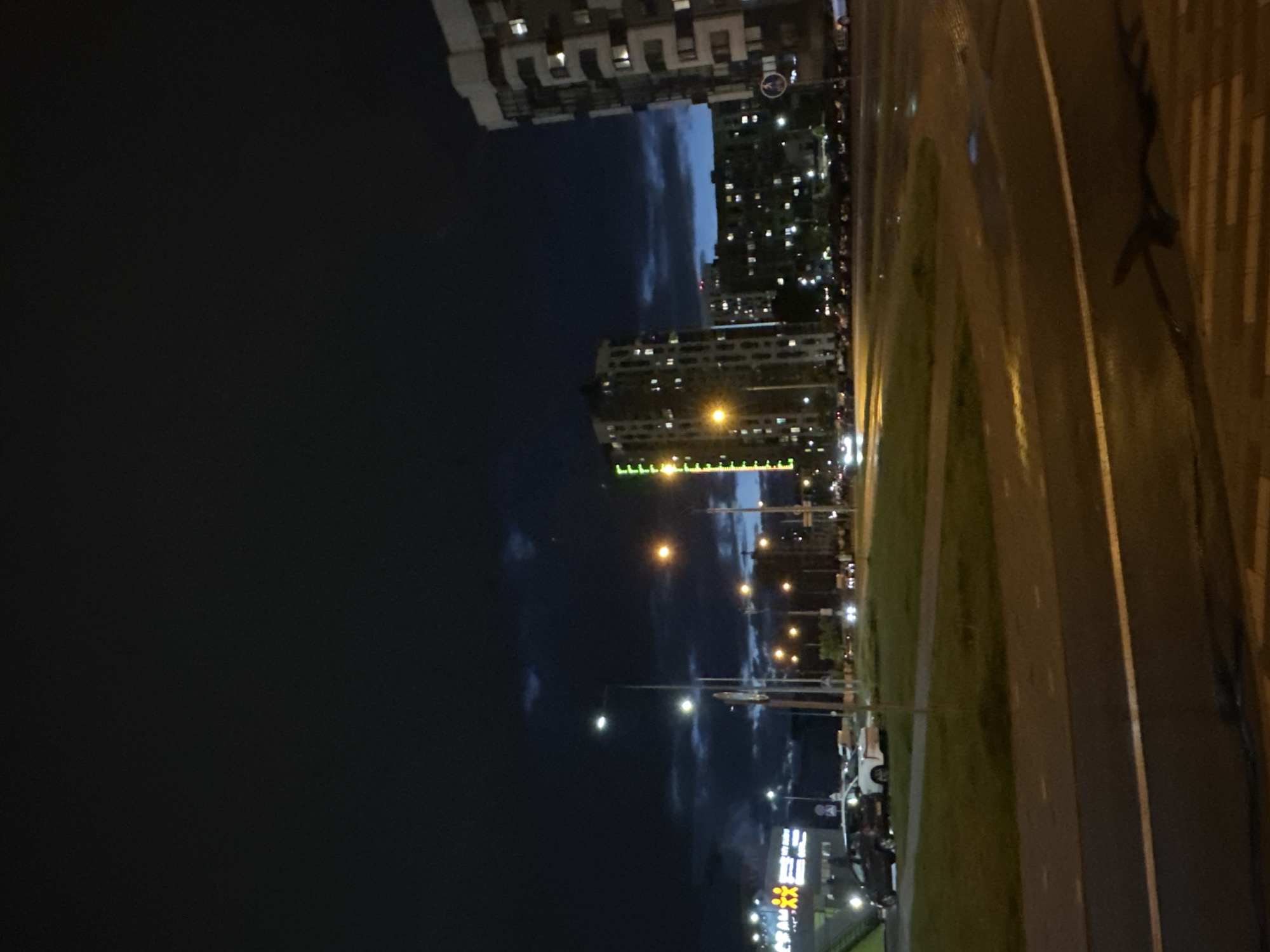 The image shows a night scene in Бараўлянскі сельскі Савет, Belarus. Tall buildings with illuminated windows are visible in the background. Streetlights illuminate the scene. A road runs along the right side of the image, with visible lane markings. A grassy area separates the road from the buildings. Parked cars are seen along the road and near the buildings. The sky is dark with patches of clouds.