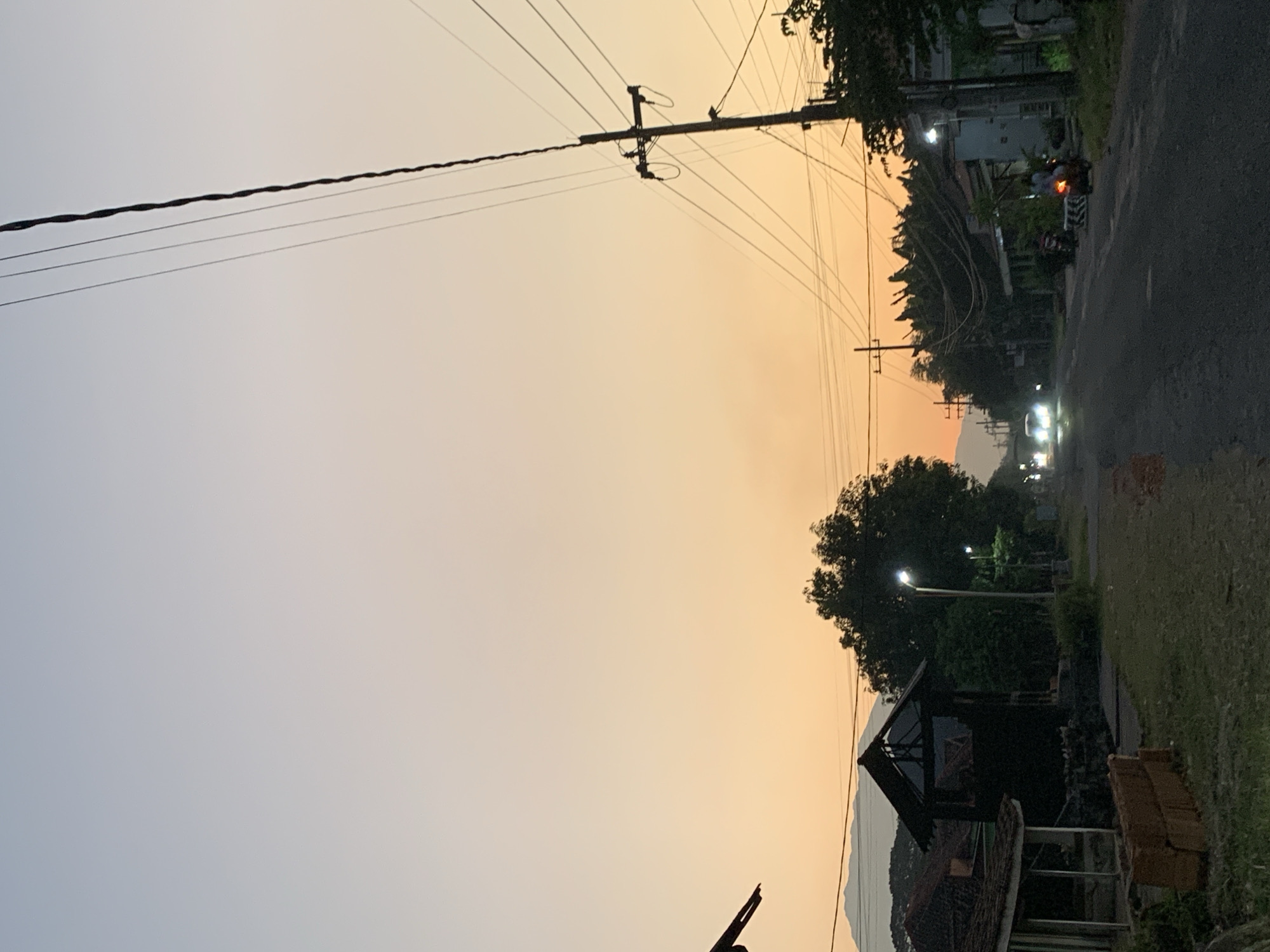 The image captures a rural or semi-urban street in Maospati, Indonesia, during either dawn or dusk. The sky exhibits a gradient, transitioning from a pale blue-grey at the top to a warm orange-yellow and reddish hue near the horizon, where distant mountains are silhouetted on the left. An asphalt road occupies the central and right lower portions of the frame, bordered by grassy verges. On the left, a stack of red bricks sits on the ground next to a path, which leads to a dark, open-sided structure and a building with a tiled roof. Numerous utility poles with a complex network of overhead power lines are visible, with a prominent pole on the far right. Streetlights are illuminated, emitting bright white light. Along the roadside, various residential or commercial buildings are interspersed with lush tropical vegetation, including trees and bushes. Two vehicles with illuminated headlights are approaching from the distant end of the road, while a person is riding a motorcycle closer to the foreground on the right, its taillight visible. Another motorcycle is parked further ahead on the right.