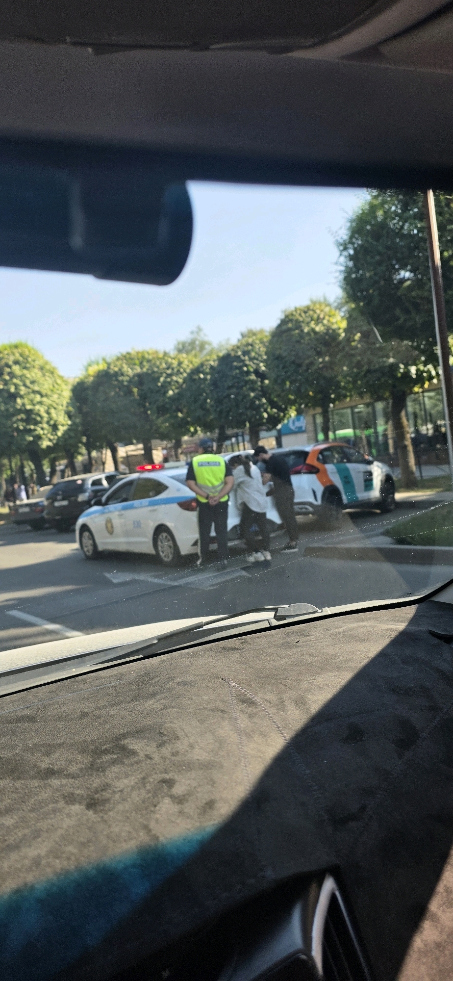 A police officer in a bright yellow vest and dark pants stands next to a white police car with blue and yellow markings. The officer has his hands behind his back. A white and orange car is parked next to the police car, and two civilians are interacting with the car. One civilian, wearing a light-colored top and dark pants, appears to be leaning into the car. Another civilian, in dark clothing, is standing beside them. The scene appears to be on a street in Almaty, Kazakhstan, with trees lining the sidewalk and buildings visible in the background. The sun is shining, casting shadows on the road.