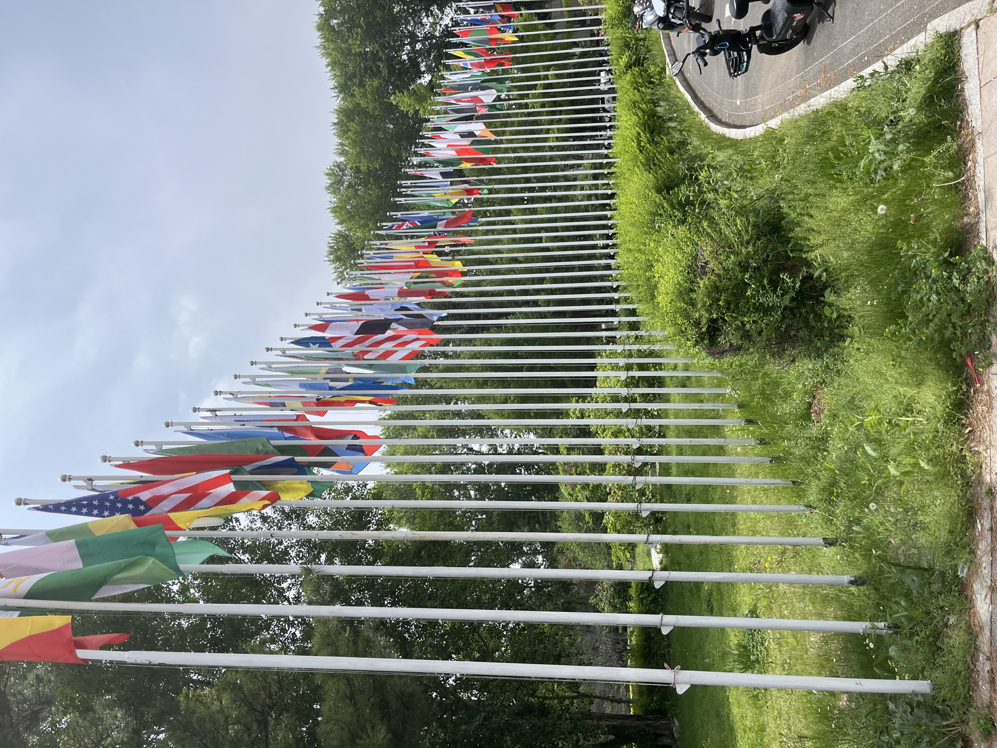 The image shows a row of flags of various countries displayed on white poles. The poles are arranged in a line, and the flags are fluttering in the wind. The background includes green trees, shrubs, and grass. There is also a paved area with two scooters parked on it. The location is implied to be Changchun City, China, based on external information.