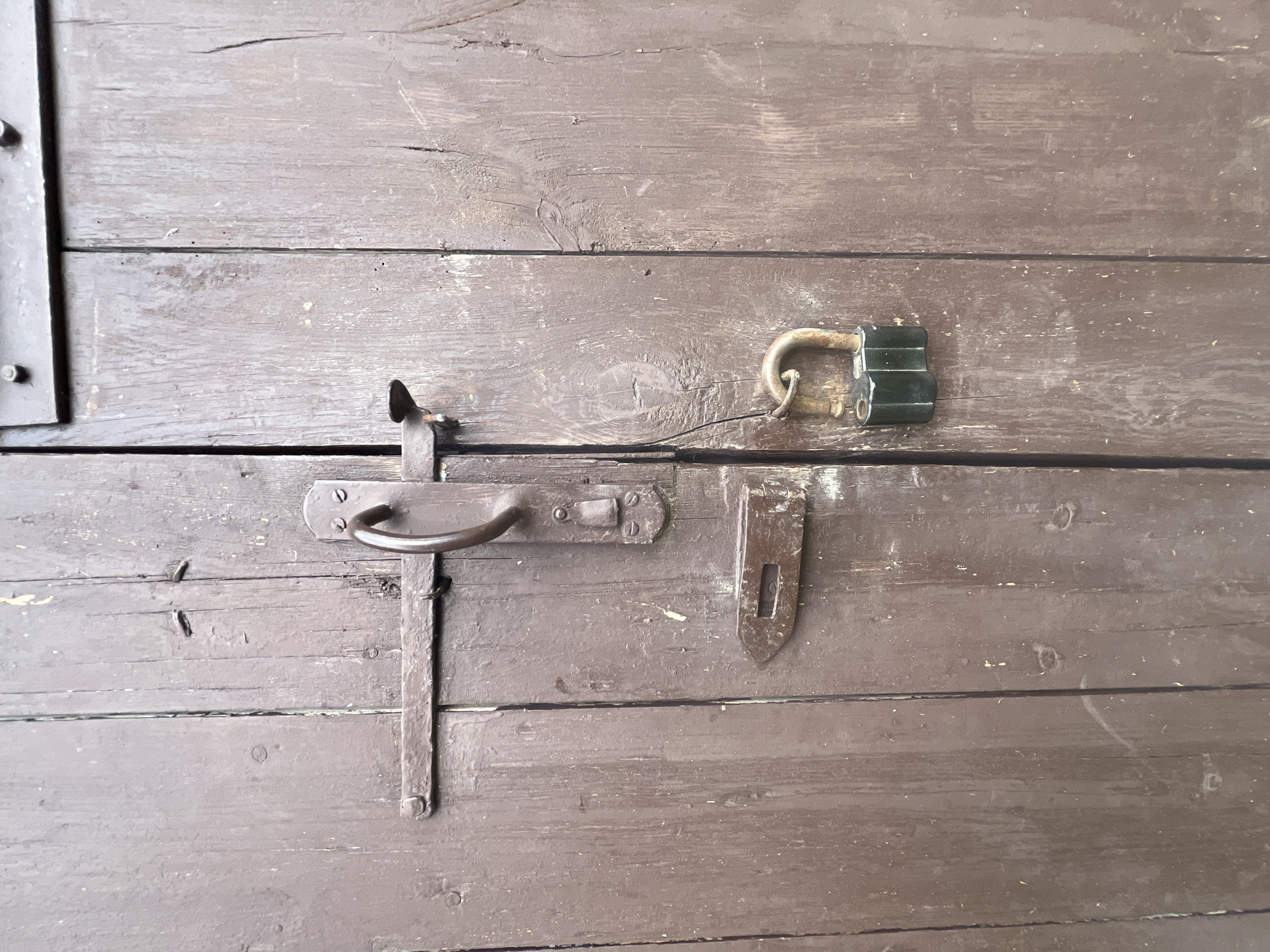 The image shows a weathered, dark brown wooden door with vertical planks. A metal latch mechanism with a handle is visible on the left side of the door. A hasp is attached to the door, and a green padlock is secured to it, indicating the door is locked. The wood appears old and worn, with some paint peeling and visible grain. The environment suggests an outdoor setting, potentially in the rural area of Бучацінскі сельскі Савет, Belarus, given the rustic nature of the door and its hardware. There are no people or animals present in the image.