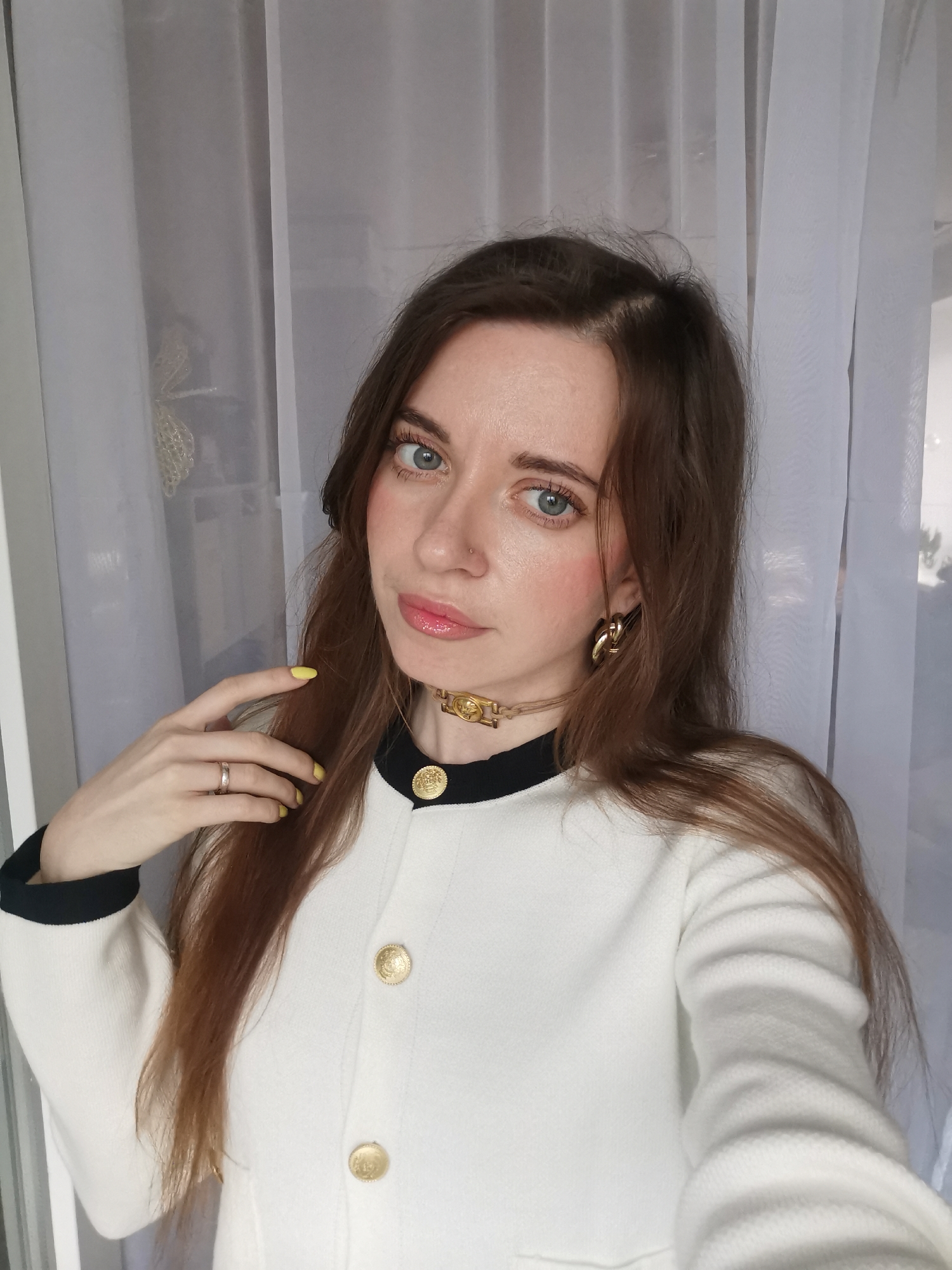 A young woman with long, straight light brown hair and bright blue eyes is captured in a close-up, chest-up portrait. She looks directly at the camera with a calm expression, her lips glossy pink and cheeks lightly blushed. A small silver nose stud is visible on her left nostril.
She is wearing a white jacket or cardigan with a black collar and black trim, adorned with three prominent gold-colored, ornate round buttons down the front. Around her neck, she wears a gold-colored choker-style necklace featuring a detailed gold emblem at the center. Large, chunky gold-colored chain-link earrings dangle from her ears. Her fingernails are painted a vibrant yellow, and a simple silver-colored ring is on the ring finger of her left hand, which is gently raised towards her hair/neck area.
The background consists of sheer white curtains, suggesting an indoor setting, likely near a window, with soft, natural light illuminating the scene. No text is visible in the image.