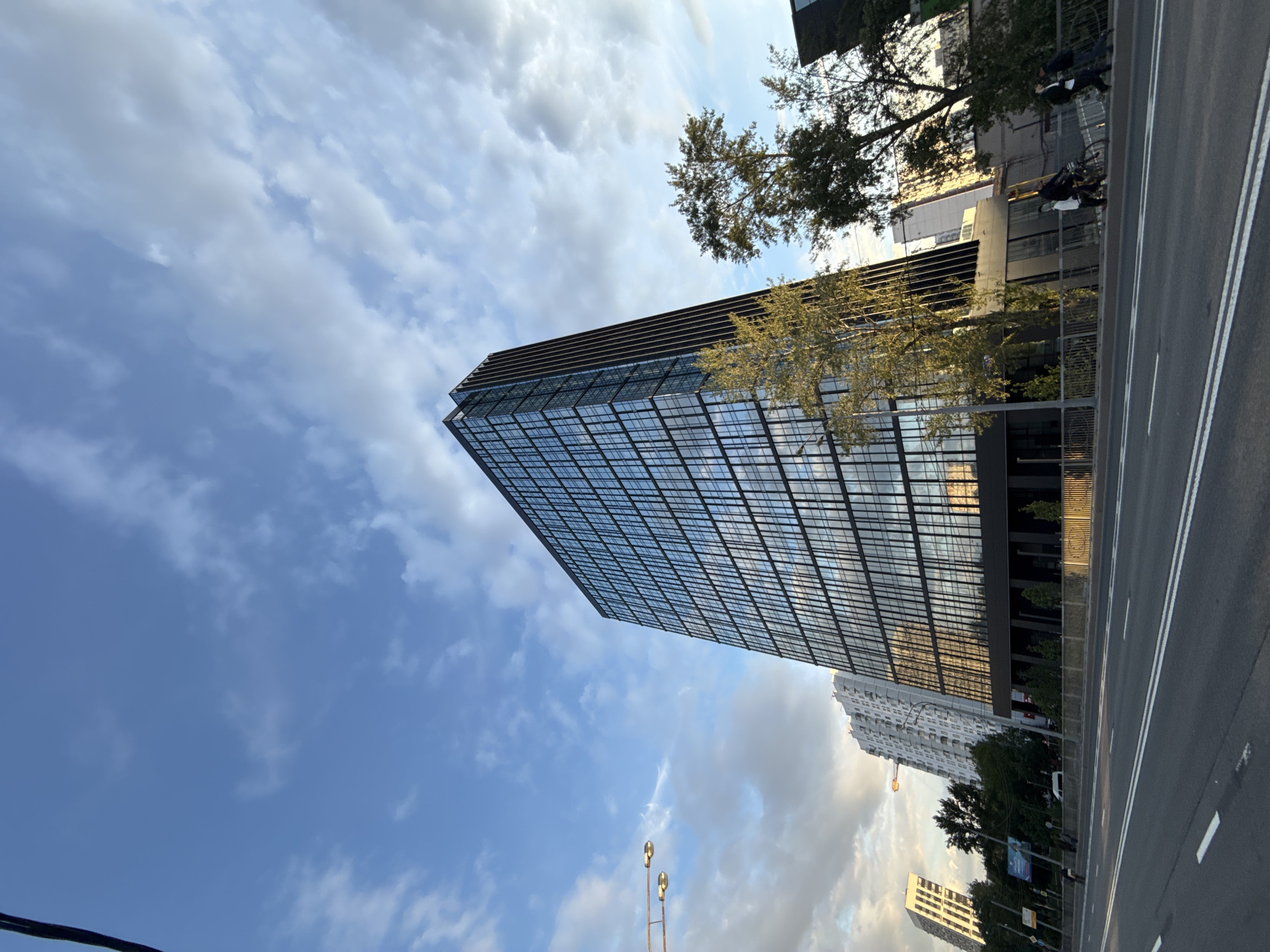 The image, correctly oriented, presents a modern urban street scene in Moscow, Russia, likely captured in the late afternoon or early evening. A prominent feature is a tall, contemporary skyscraper on the left, distinguished by its dark facade and a grid pattern of numerous reflective glass windows that mirror the sky and surrounding environment. Several trees with green foliage are visible, some partially obscuring the lower and middle sections of the main building, and others lining the street.
To the right of the building, a wide, multi-lane asphalt road stretches into the distance, clearly marked with white lane lines. A robust metal barrier runs along the side of the road, separating it from the pedestrian area and the buildings. In the background, further down the street, a lighter-colored high-rise building, possibly residential, can be seen, with a construction crane next to it. Another smaller, white building is also visible in the distance.
The sky occupies a significant portion of the upper frame, showcasing patches of clear blue interspersed with scattered, fluffy white clouds. The lighting suggests either a low sun or an overcast period, with reflections visible on the glass surfaces.
Several individuals are present in the scene: a figure is walking on the sidewalk, and another person appears to be riding a bicycle near the road. Two multi-lamp streetlights rise from the lower foreground. In the middle distance, a blue advertising billboard is discernible, but any text on it is not legible due to the distance. The scene conveys the character of an active city environment.