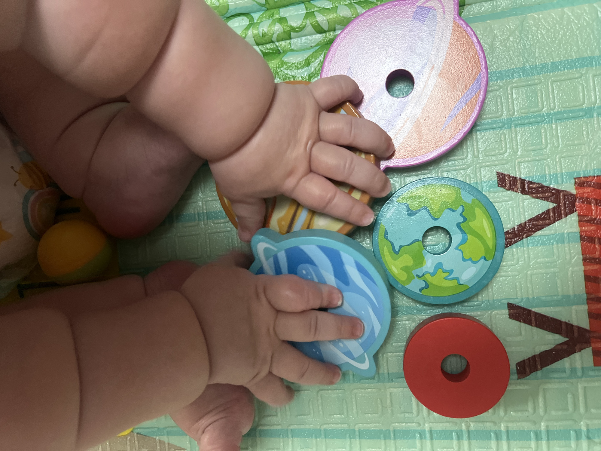 This is a close-up, overhead shot of a baby's hands interacting with colorful, planet-themed wooden toys on a green play mat. The baby's chubby arms and hands are prominent in the foreground. The toys include a blue planet with white swirls, a green and blue earth-like planet, a pink and orange planet, and a red ring. The play mat has a grid pattern. The background is slightly blurred but shows parts of the baby's legs and possibly other soft toys. The lighting is bright, suggesting an indoor environment. The scene is devoid of any discernible urban elements, making it impossible to determine a location in the city of Unknown, Unknown.