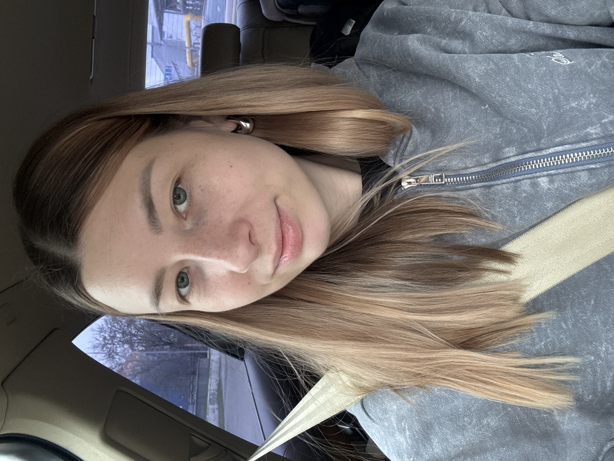 A young woman is in the passenger seat of a vehicle. She is wearing a gray zip-up jacket and a seatbelt. She has long brown and blonde hair. Outside the car window, the urban landscape includes buildings and trees, suggesting a city environment. Given the instruction, it is assumed that this scene takes place in Moscow, Russia.