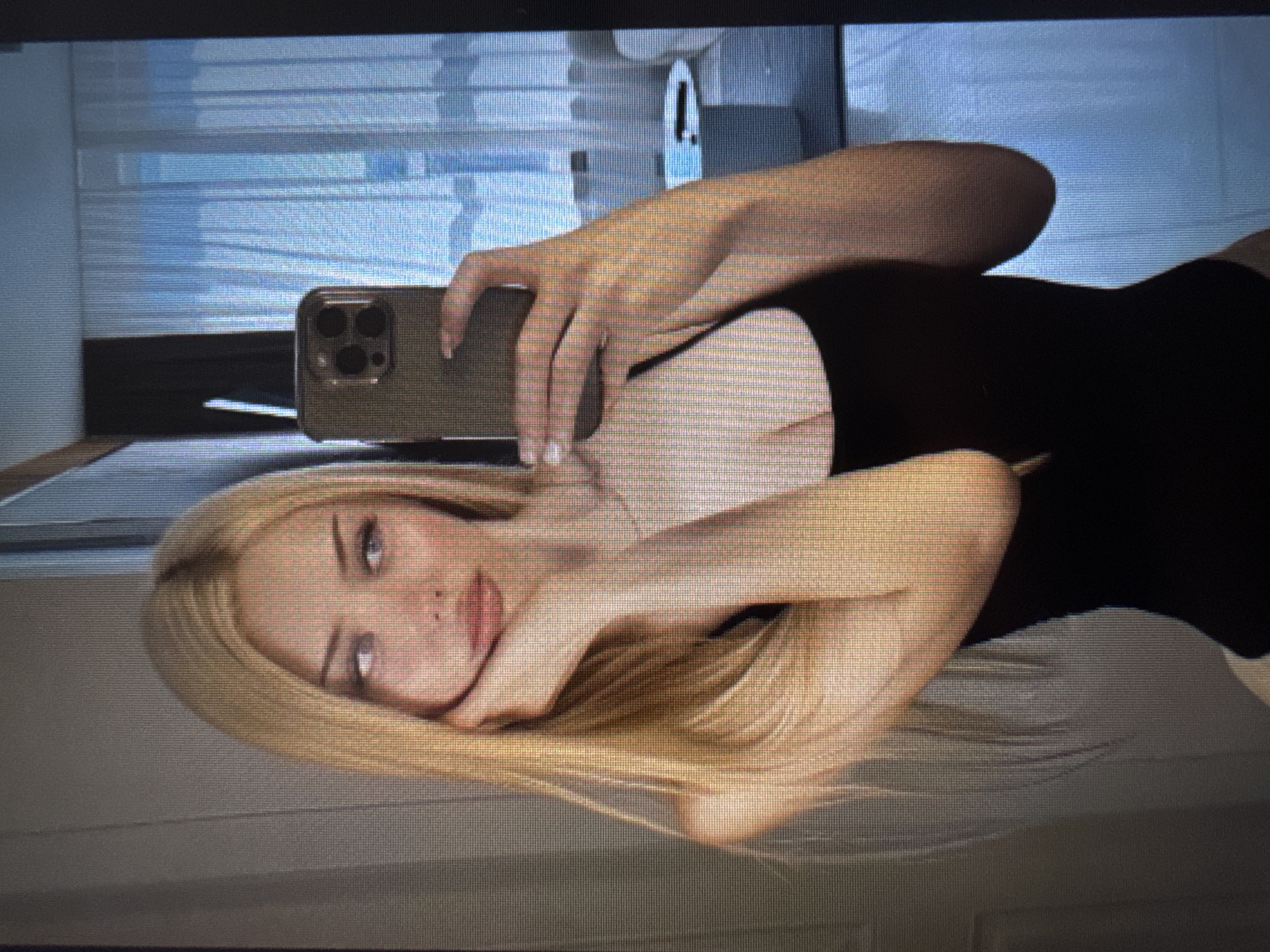 The image is a portrait of a fair-skinned woman with long, straight blonde hair and light-colored eyes, taken at a slightly Dutch angle and rotated 90 degrees clockwise. She is wearing a dark, possibly black, top and has a neutral expression, looking directly towards the camera.
She is holding a smartphone, identifiable as an iPhone due to its triple-lens camera module, encased in a dark-colored protective cover. Her fingers are visible wrapped around the device as she takes a selfie or self-portrait.
The background shows horizontal window blinds, indicating an indoor setting with diffused light filtering through, suggesting daytime. The image itself appears to be a photograph of a screen, exhibiting noticeable pixelation and scan lines. There is no visible text in the image.