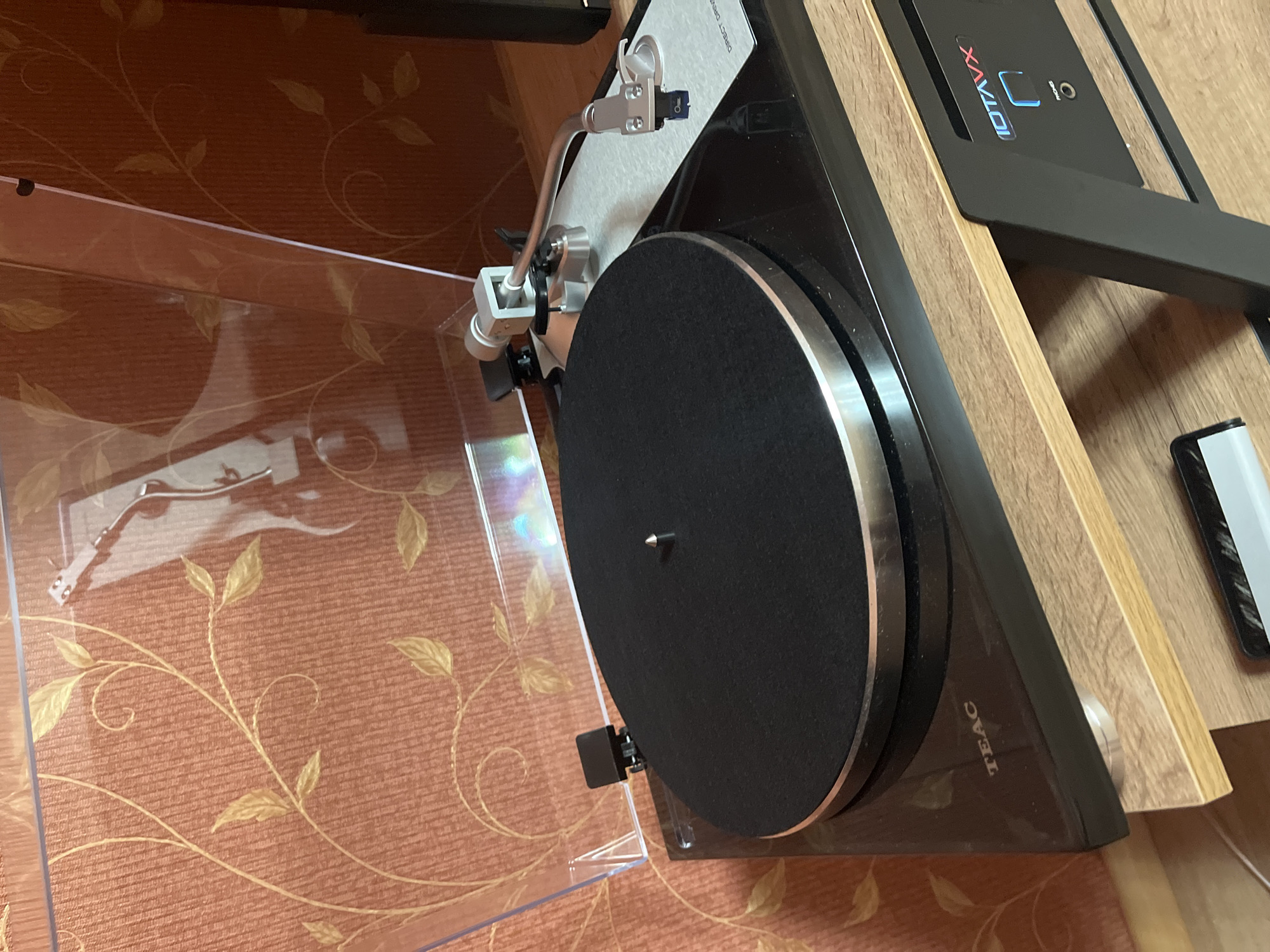 The image shows a close-up, eye-level view of a black TEAC direct drive turntable. The turntable has a silver trim around its black platter, which has a textured surface. A tonearm with a silver finish is positioned above the platter. The turntable is placed on a light wood shelf unit. In the background, there is an orange textured wallpaper with a subtle gold floral pattern. To the right of the turntable, a black electronic device with a blue illuminated display that reads "IOTAVX" is visible. The scene appears to be indoors, likely in a domestic setting. There are no people or animals present in the image, and no specific location in the city of Яново-Грушевский, Russia can be identified from the visual information provided.