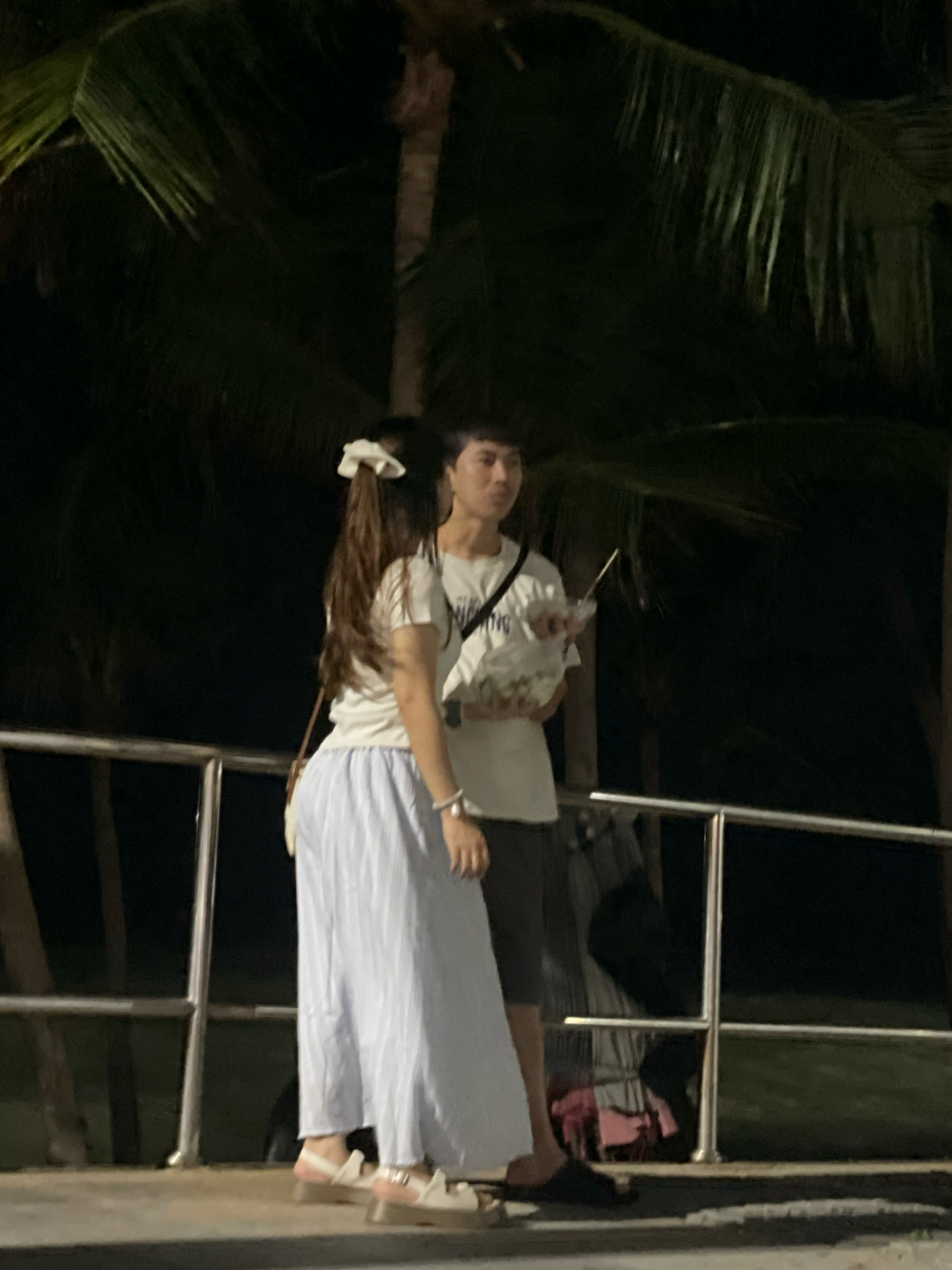 A young man and woman are standing side-by-side at night, outdoors, with palm trees in the background. The man, on the right, is wearing a white t-shirt and dark shorts, and holding a clear plastic bag with a straw. The woman, on the left, has long brown hair with a white scrunchie, is wearing a white t-shirt and a long, light blue pleated skirt, and a white bracelet on her wrist. They are standing near a metal railing. The overall scene suggests a casual evening setting, possibly a promenade or park area in Pattaya City, Thailand.