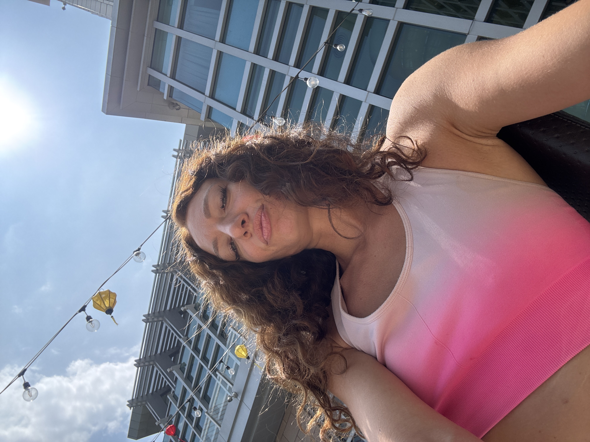 A woman with long curly brown hair is posing for a selfie. She is wearing a pink and white ombre sports bra. Behind her is a modern building with glass windows and a string of lights and lanterns. The location is indeterminate. Đà Nẵng, Vietnam, cannot be determined from the image.