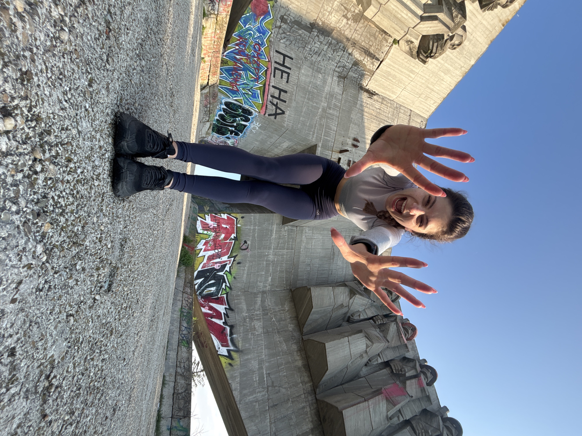 This vertical shot, taken from a low angle, features a young woman playfully posing in front of a large concrete monument or structure, with a clear blue sky in the background. The scene is set in Varna, Bulgaria.
The woman, with dark hair tied back, wears a light grey long-sleeved top, dark blue leggings, and black athletic shoes. She is looking directly at the camera with a wide, joyful smile. Her hands are extended towards the camera with fingers spread, creating a forced perspective effect that makes them appear large and close to the viewer. Her fingernails are painted a light red/pink.
The imposing concrete walls behind her are characteristic of brutalist architecture, featuring geometric and blocky shapes, possibly depicting abstract figures or reliefs. These structures are extensively covered in vibrant graffiti, including stylized lettering in various colors like blue, green, red, and black. The phrase "HE HA" is clearly visible in black text on one of the concrete surfaces. The ground where the woman stands is a rough, pebbled concrete texture.
The lighting suggests a bright, sunny day, with strong natural light casting shadows on the concrete, indicating either morning or late afternoon. The overall atmosphere is cheerful and dynamic, reflecting the woman's energetic pose and happy expression amidst the rugged, graffiti-adorned urban landscape.