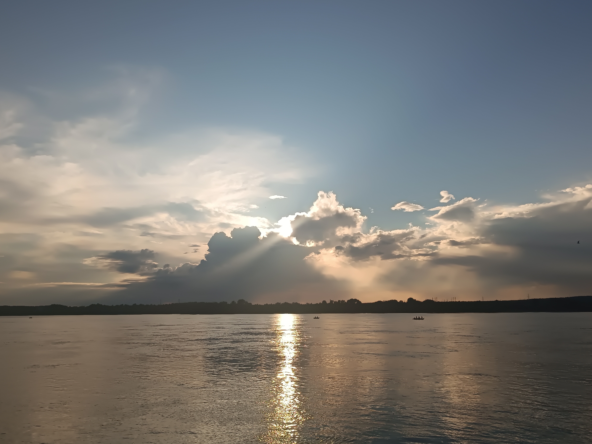 This image depicts a wide, calm river with a reflective surface, likely the Kama River in Chaikovsky, Russia. The sun is partially obscured by dramatic, illuminated clouds on the horizon, casting a golden sheen across the water. A dark, silhouetted treeline borders the far bank of the river, dotted with what appear to be distant structures. Three small boats, each with at least two occupants, are visible on the water, one closer to the left bank and two closer to the center. The sky transitions from a pale blue at the top to a softer, cloud-filled expanse near the horizon. The overall atmosphere is serene, suggesting either early morning or late afternoon.