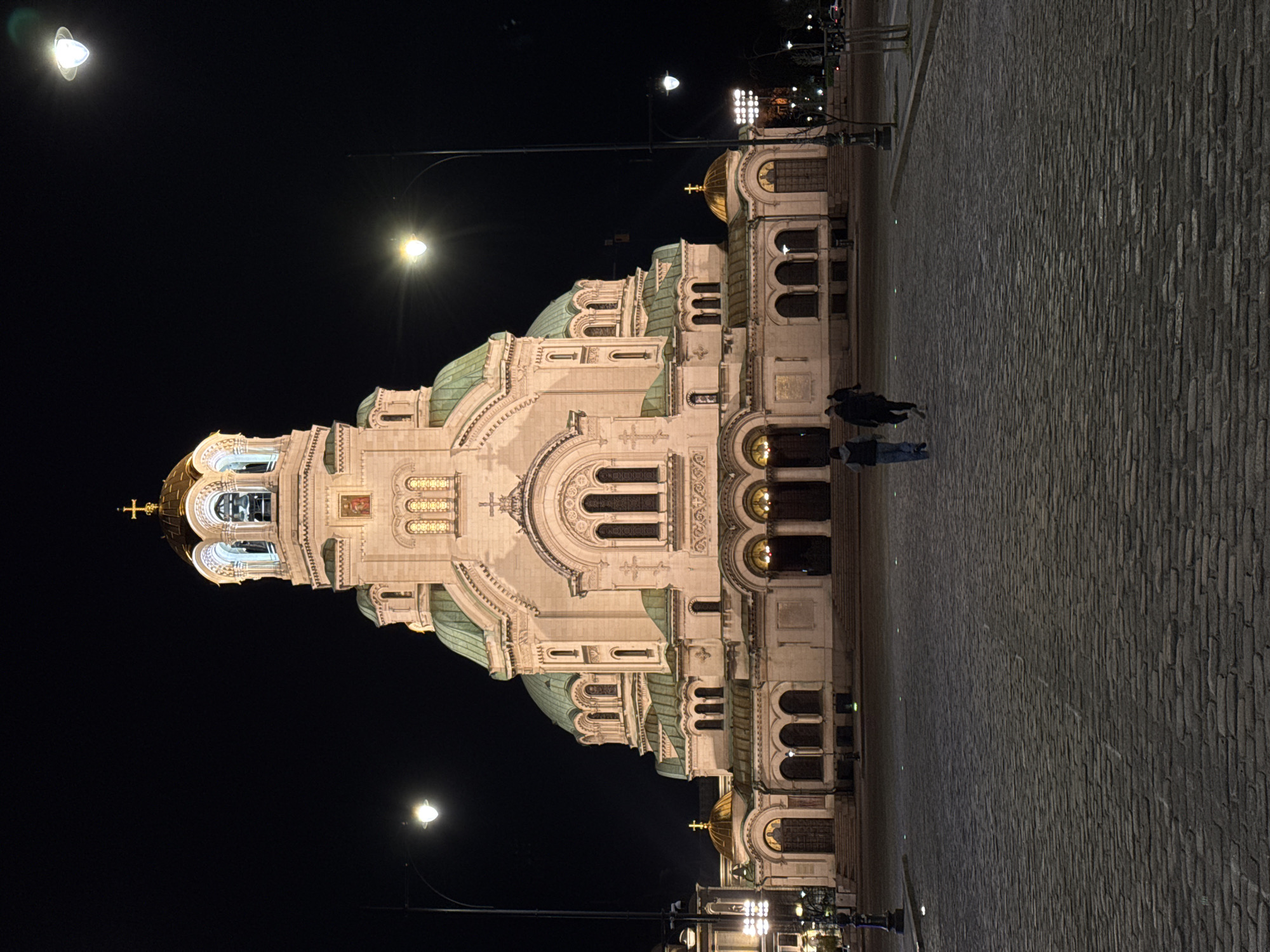 The image shows the Alexander Nevsky Cathedral in Sofia, Bulgaria, at night. The cathedral is illuminated and dominates the right half of the image. A cobblestone pavement stretches from the bottom edge of the image towards the cathedral. Two people are walking on the pavement towards the cathedral. Several streetlights are visible, illuminating the scene. The sky is dark and clear.