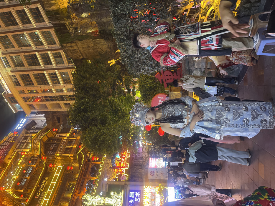 The image captures a nighttime urban scene in the Yuzhong District of Chongqing, China, prominently featuring cultural performers against a backdrop of traditional and modern architecture. In the foreground, center-left, a female stands facing slightly right, adorned in an elaborate light blue and silver traditional ethnic costume. She wears a tall, ornate silver headdress, multiple layered silver necklaces, a silver-embellished top and skirt, and her hair is styled in long braids. To her right, a male stands on a small blue plastic stool, dressed in a red, beige, and multi-patterned traditional costume with fur trim, holding a small drum near his chest.
Behind the performers, numerous pedestrians are visible on a paved street, moving through a brightly lit area. In the background on the left, a large, multi-story traditional Chinese building with tiered roofs and balconies is extensively illuminated with warm yellow strip lights and numerous red lanterns. To the right of this, a modern high-rise building with multiple windows ascends a natural rock face and is also illuminated. Green trees and foliage are present between the foreground subjects and the background buildings. Illuminated signs, including one for "CHAGEE" and others with Chinese characters, are visible in the midground. The scene is brightly illuminated by artificial lighting, indicating nighttime.