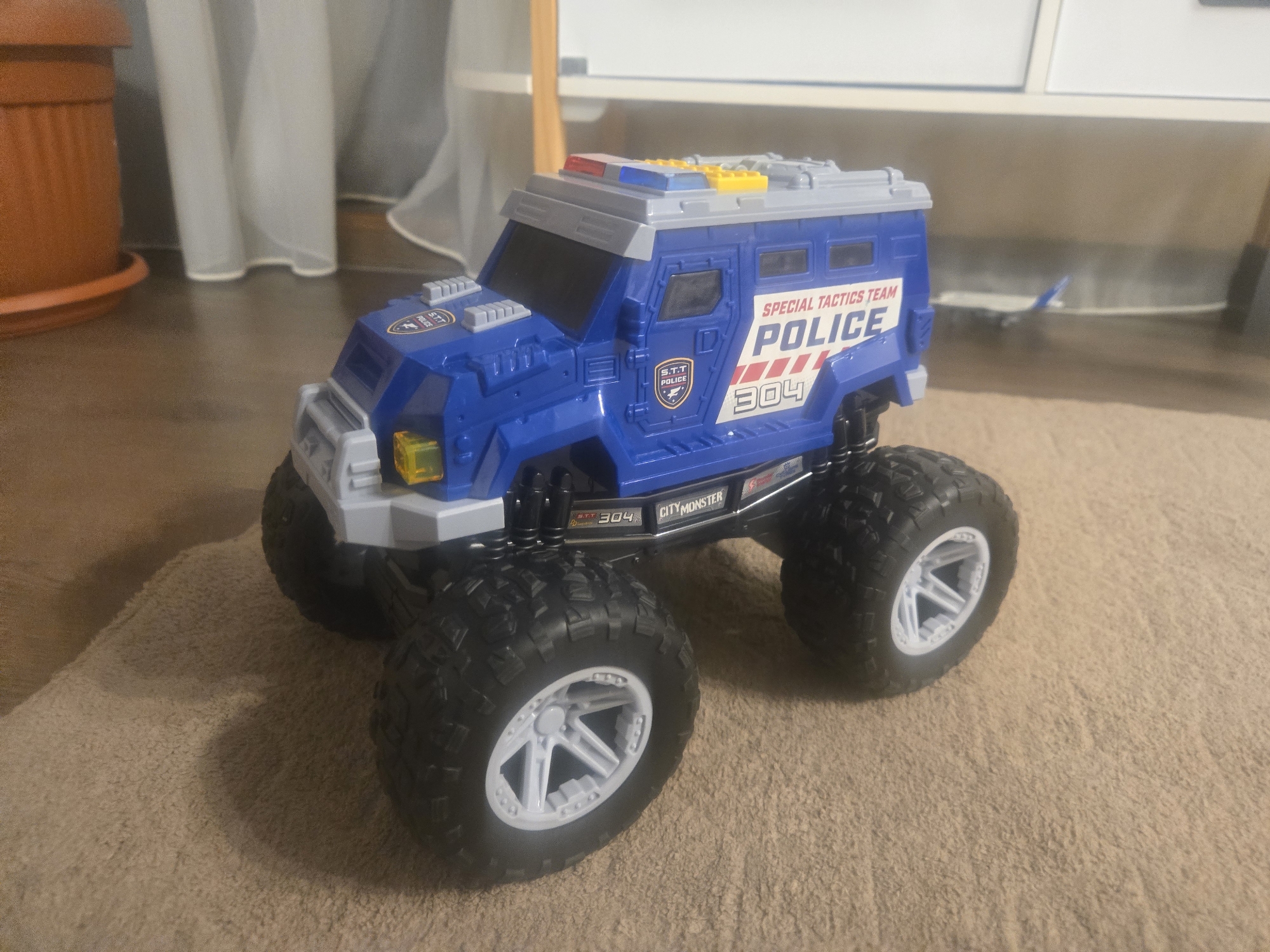 A blue and grey toy police monster truck sits on a tan carpet. The toy truck has large black tires with white rims, and its body is primarily blue with "SPECIAL TACTICS TEAM POLICE 304" printed on the side. The top of the truck is grey with colored lights. In the background, a brown flower pot and white curtains are visible, along with a white cabinet. The location appears to be indoors, possibly a home.
