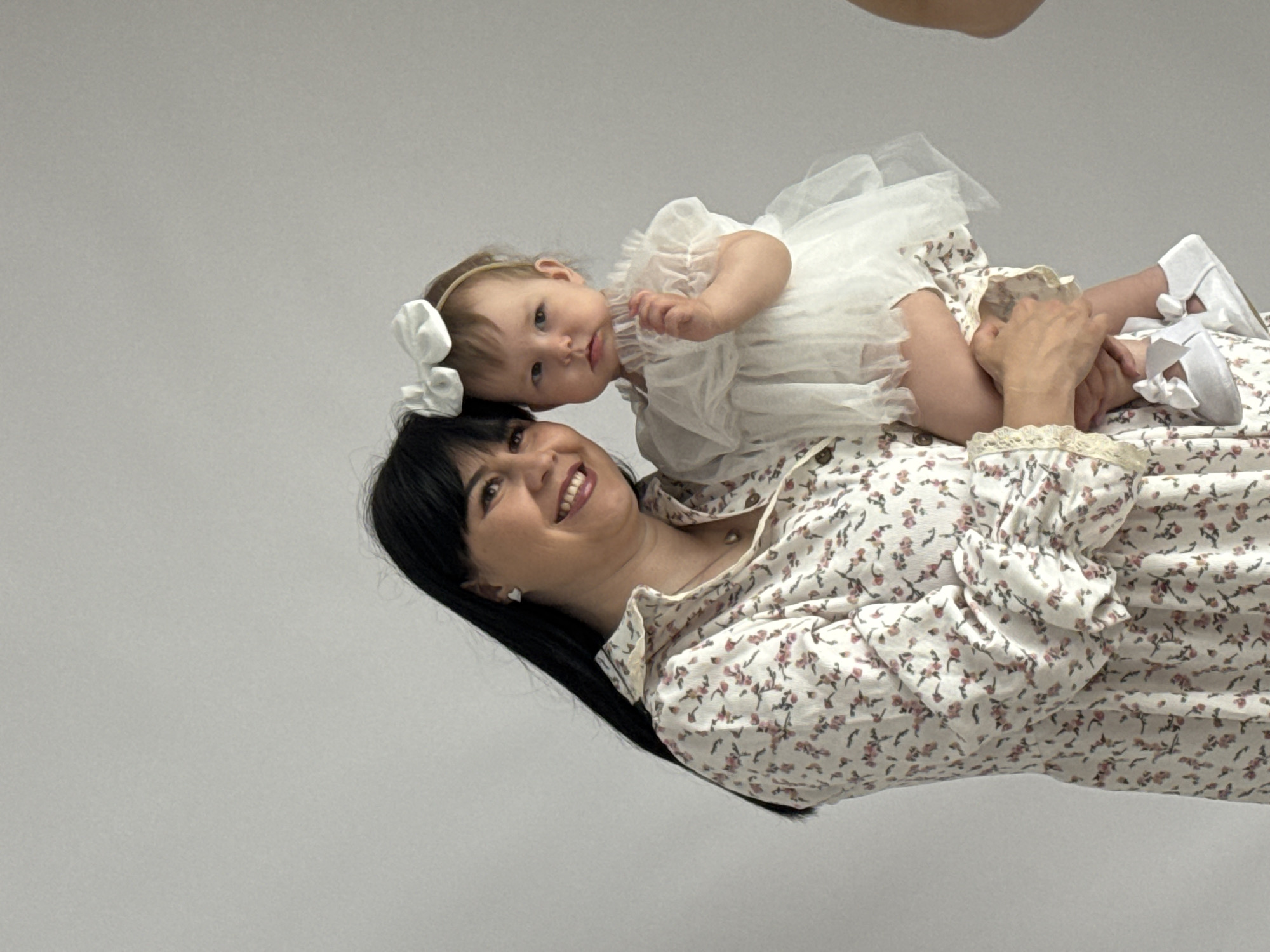 A medium shot depicts a woman and baby. The woman is lying on her back, wearing a floral-patterned blouse. She has black hair, a fair complexion, and is smiling. Atop the woman's torso lies the baby, wearing a white dress and headband with a bow. The baby's eyes are open. A hand is visible in the top right corner of the frame. The background is a plain grey. The scene appears to be set indoors, possibly in a studio setting. There is no indication that this image was taken in Izhevsk, Russia.