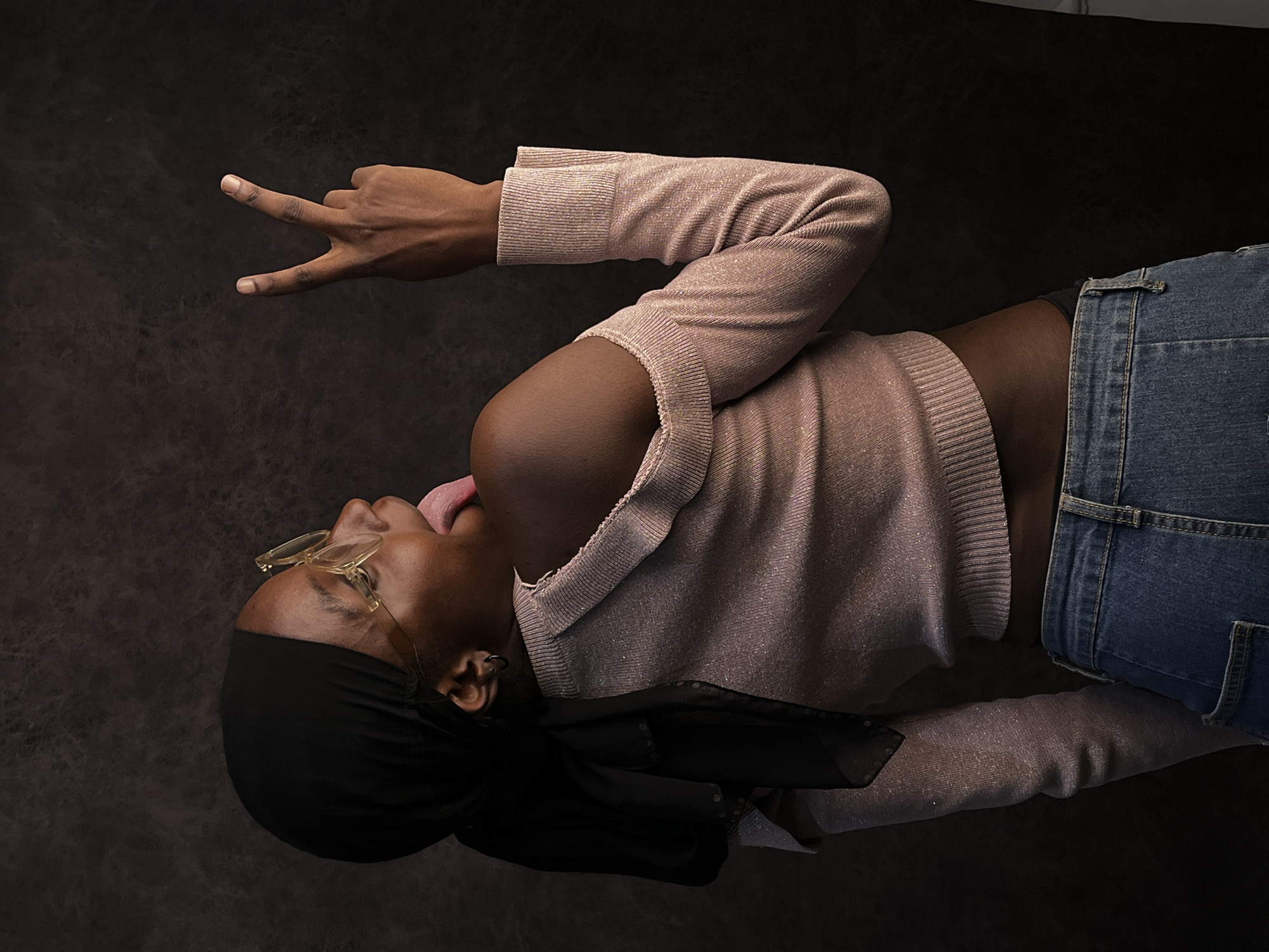 A dark-skinned person wearing glasses and a black head covering lies on a dark surface. The person wears a light pink long-sleeved top and blue jeans. Their tongue is sticking out, and they are holding up their hand, making a peace sign. The background is a mottled dark gray/black. The location of Jos, Nigeria, cannot be determined from the image.