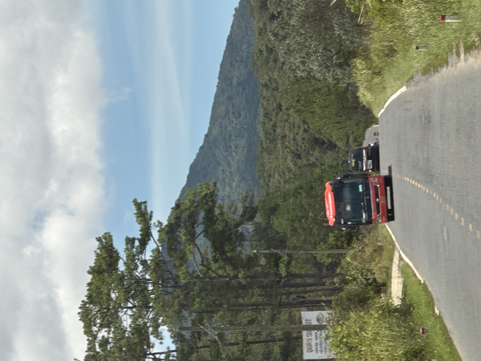 The image depicts a paved road winding through a landscape characterized by dense green trees and rolling hills under a partly cloudy sky. A bright red bus is in the foreground, traveling away from the camera. To the left of the road, there is a white sign with text, partially obscured by foliage. Further down the road, a dark-colored van and a person on a scooter are visible in the distance. The road is lined with grass and small red and white markers. The overall scene suggests a rural or mountainous area. While the prompt requests information about the location in the city of Đưng K'Si, Vietnam, this specific detail cannot be confirmed solely from the visual information in the image.