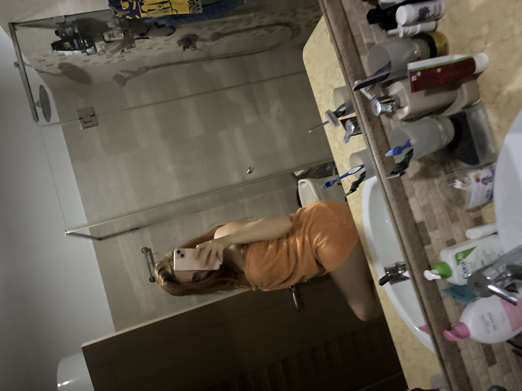In a brightly lit bathroom, a woman with long, auburn hair, wrapped in an orange towel, takes a selfie in a mirror. She is positioned on the left side of the frame, her body angled towards the mirror as she holds up a pink smartphone to capture her reflection.
The bathroom features a modern shower enclosure with glass doors and chrome fixtures. On a shelf inside the shower, several bottles of toiletries are visible. To the right of the shower, a yellow and blue patterned towel hangs.
The vanity area, visible in the foreground, is cluttered with various bathroom essentials. A white sink is present, with a chrome faucet. On the countertop, there are several toothbrush holders containing toothbrushes, a bottle of CeraVe lotion, and other personal care items. The walls are tiled with a mosaic pattern. The overall environment suggests a residential bathroom.