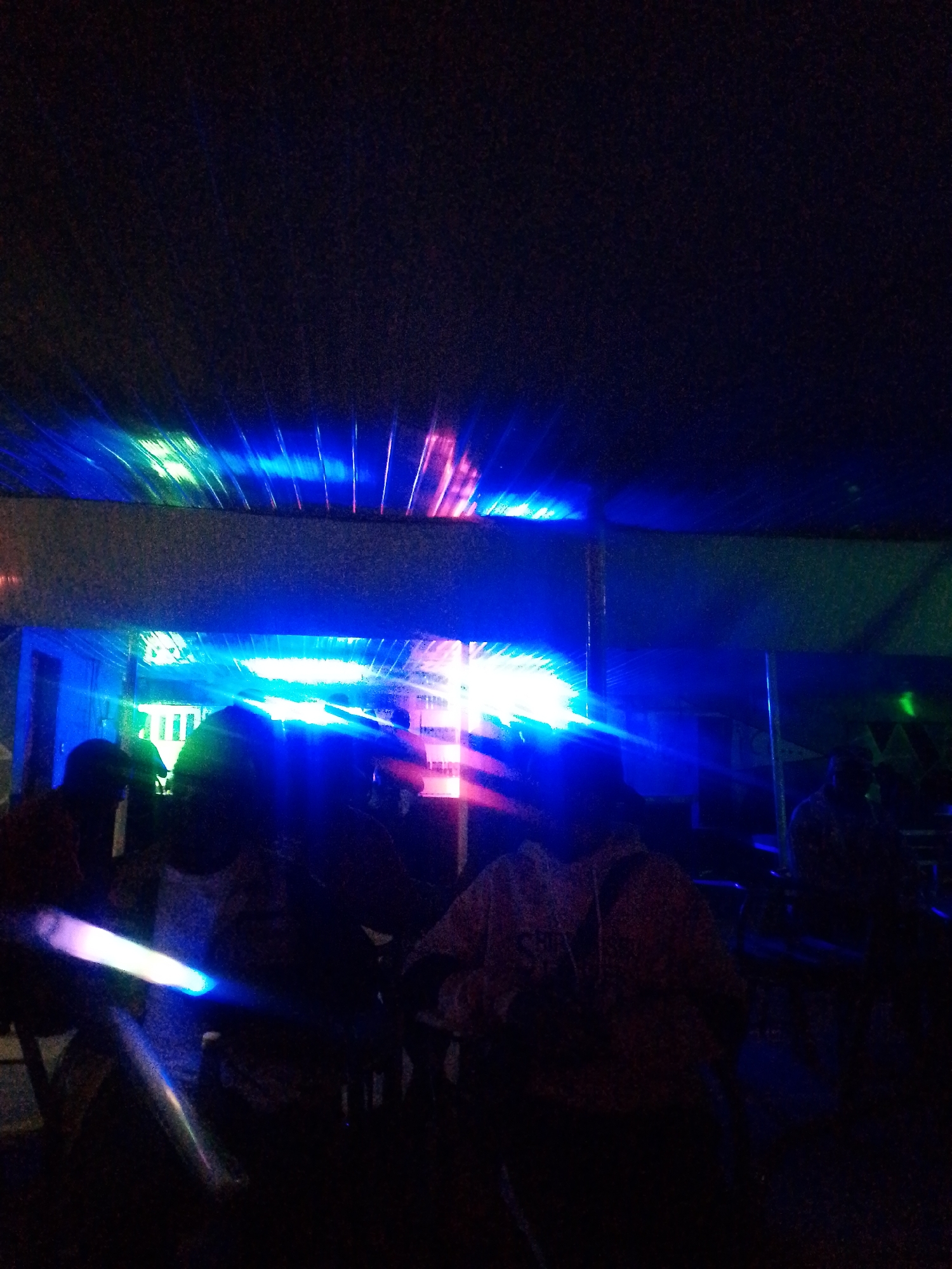 The image captures a dimly lit scene, likely a night time event or venue, characterized by vibrant blue and pink light beams. The lighting emanates from above, creating streaks of color across a dark background. Silhouettes of several people are visible, suggesting they are observing or participating in the illuminated display. The environment appears to be indoors, with elements that could indicate a stage, ceiling, or a decorated area. The focus is on the interplay of light and shadow, with the human figures serving as secondary elements within the broader atmospheric composition.