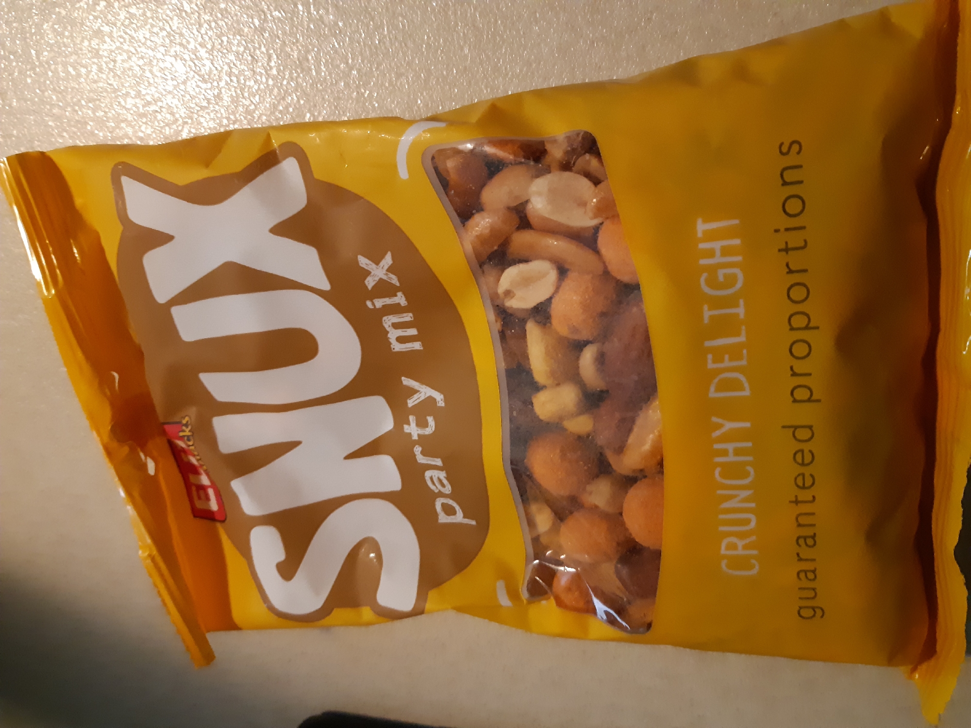 The image shows a yellow bag of "XUNS party mix" with the description "CRUNCHY DELIGHT guaranteed proportions." The bag is partially filled with a mix of nuts and other crunchy elements. The background is a neutral, lightly textured surface. There is no indication of a specific location in Plovdiv, Bulgaria, or any other city.