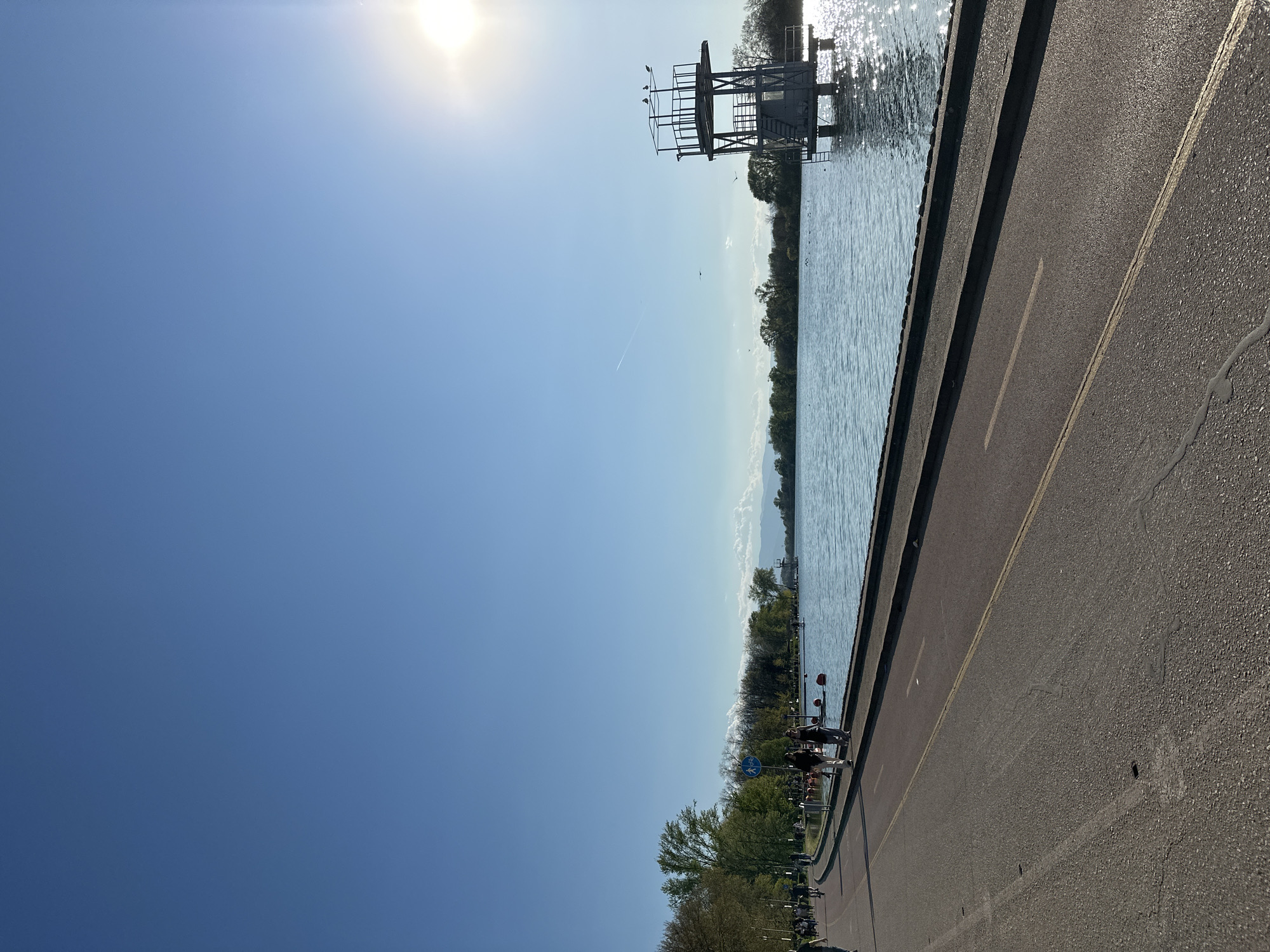 The image captures a sunny day scene near the Rowing Canal in Plovdiv, Bulgaria. The frame is divided between a clear, light blue sky and the canal itself. In the background, a metal diving platform is visible, standing in the water. Along the right side of the frame, a paved path runs adjacent to the canal, featuring yellow lines marking its lanes. Trees line the edge of the path, and people can be seen along the path. A blue bicycle sign is also visible along the side of the path.