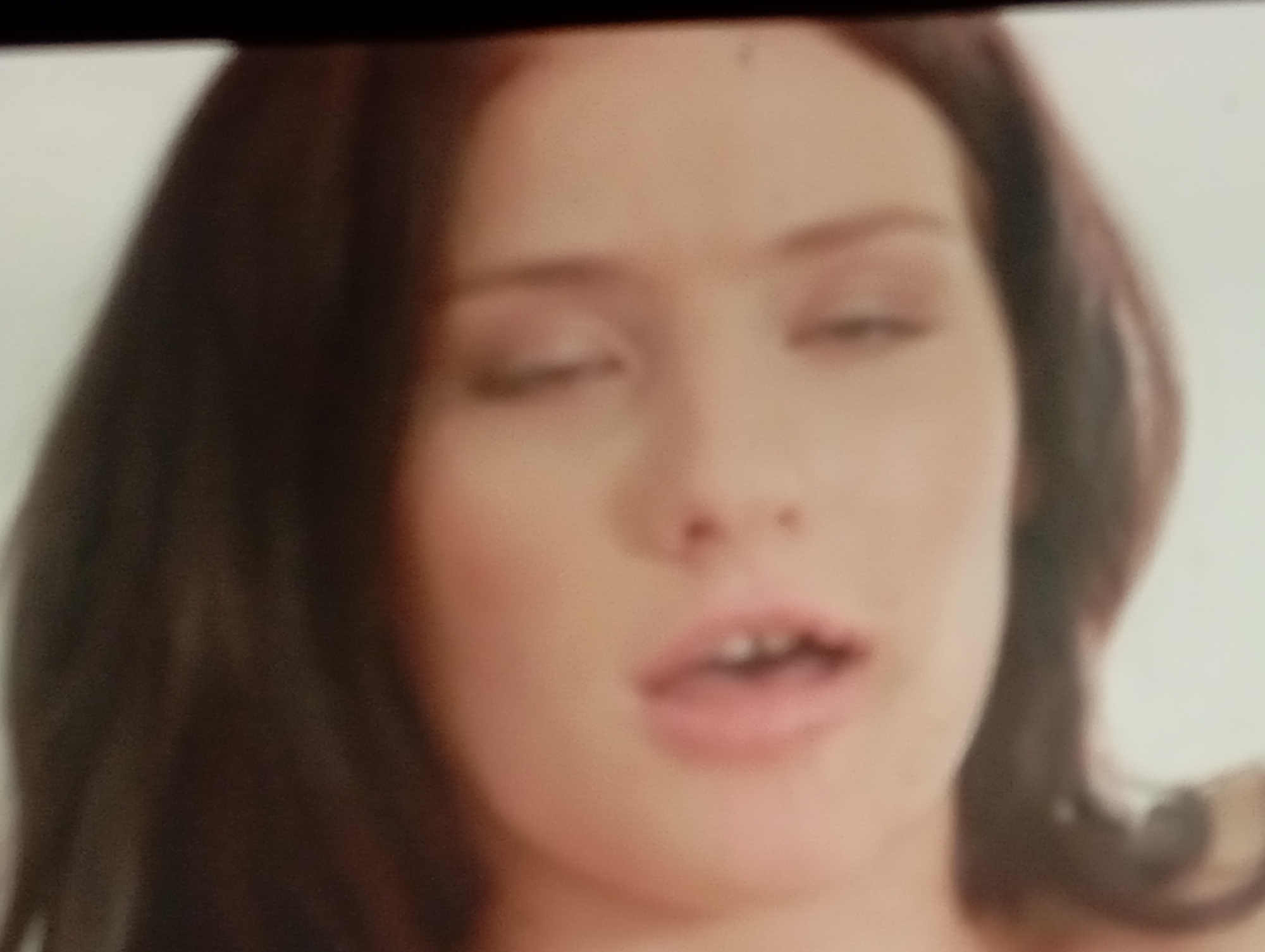 The media file is a close-up, out-of-focus shot of a woman's face. She has brown hair and is looking downwards with her eyes partially closed. Her mouth is slightly open, revealing some teeth. The lighting is soft and appears to be coming from the front, creating a gentle illumination on her skin. The background is a plain, light color, suggesting a studio setting or a neutral indoor environment. No text is visible in the image. The overall impression is one of a candid or intimate moment captured. The image's low resolution and blurriness obscure finer details and make it difficult to discern specific emotions or activities beyond the facial expression. The provided location context of Sofiivska Borshchahivka, Ukraine, does not offer any visual cues within the image itself.