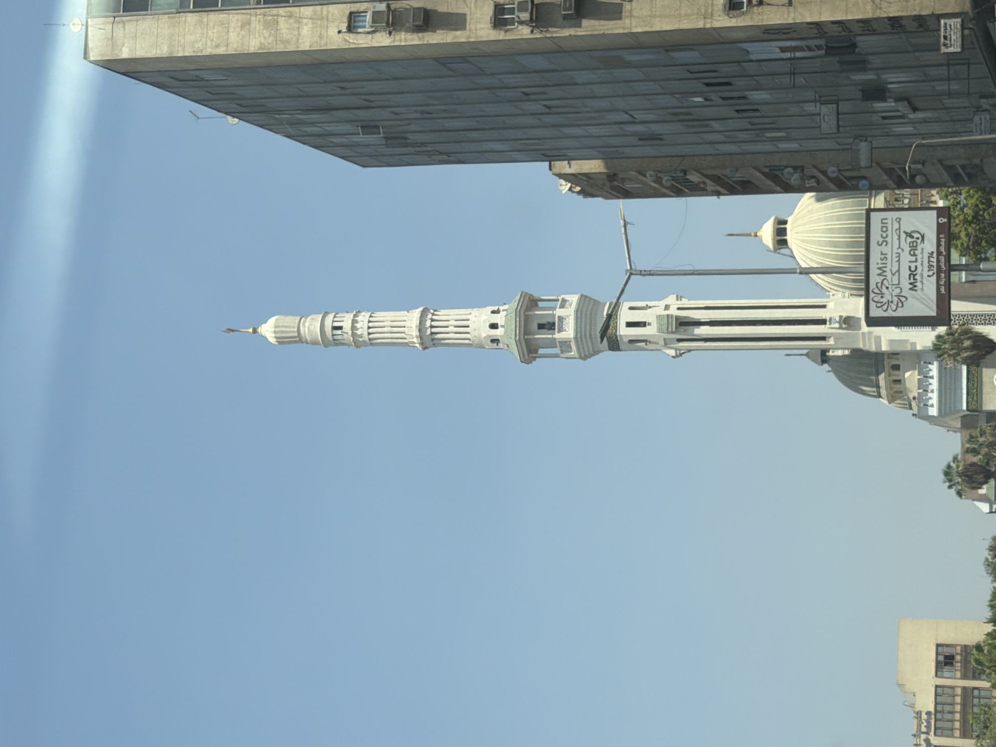 The image captures a street-level view in Cairo, Egypt, featuring a prominent mosque minaret and a large apartment building. The minaret, a tall, slender structure with intricate white latticework and green accents, is the central focal point on the left side of the frame. Adjacent to it, on the right, is a tall, gray, multi-story building with numerous windows and visible air conditioning units on its exterior. In the mid-ground, a large dome of a mosque is visible, and a billboard displaying "Misr Scan MRC LAB" with Arabic text and phone numbers is situated in front of it. The sky is a clear, bright blue with a faint white streak of light, possibly a contrail or atmospheric phenomenon. The scene depicts an urban environment with a mix of religious and residential architecture under daylight conditions.