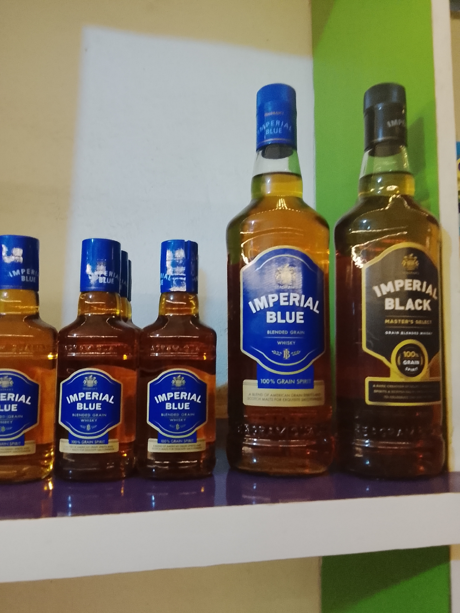 The image features five bottles of whisky neatly arranged on a dark purple shelf. To the left, there are three smaller bottles of "IMPERIAL BLUE BLENDED GRAIN WHISKY," each with a distinctive blue label and a matching blue cap, and the text "100% GRAIN SPIRIT" is visible. Adjacent to these, a larger bottle of the same "IMPERIAL BLUE BLENDED GRAIN WHISKY" stands with its blue label clearly displaying "BLENDED GRAIN WHISKY," "100% GRAIN SPIRIT," and "A BLEND OF AMERICAN GRAIN DISTILLATES AND SCOTCH MALTS FOR EXQUISITE SMOOTHNESS." On the far right, a large bottle of "IMPERIAL BLACK MASTER'S SELECT GRAIN BLENDED WHISKY" is present, featuring a dark label with yellow accents, a black cap, and text indicating "100% GRAIN Spirit" and "A RARE CREATION OF FINE INDIAN GRAIN SPIRITS & SELECTED SCOTCH MALTS FOR AN EXCLUSIVE BLEND." All bottles contain an amber-colored liquid. The background consists of a plain wall, transitioning from a light beige or off-white on the left to a bright green on the right, suggesting an indoor setting with artificial lighting. The scene appears to be a display of these alcoholic products, possibly in a store or a home collection, in Jos, Nigeria.