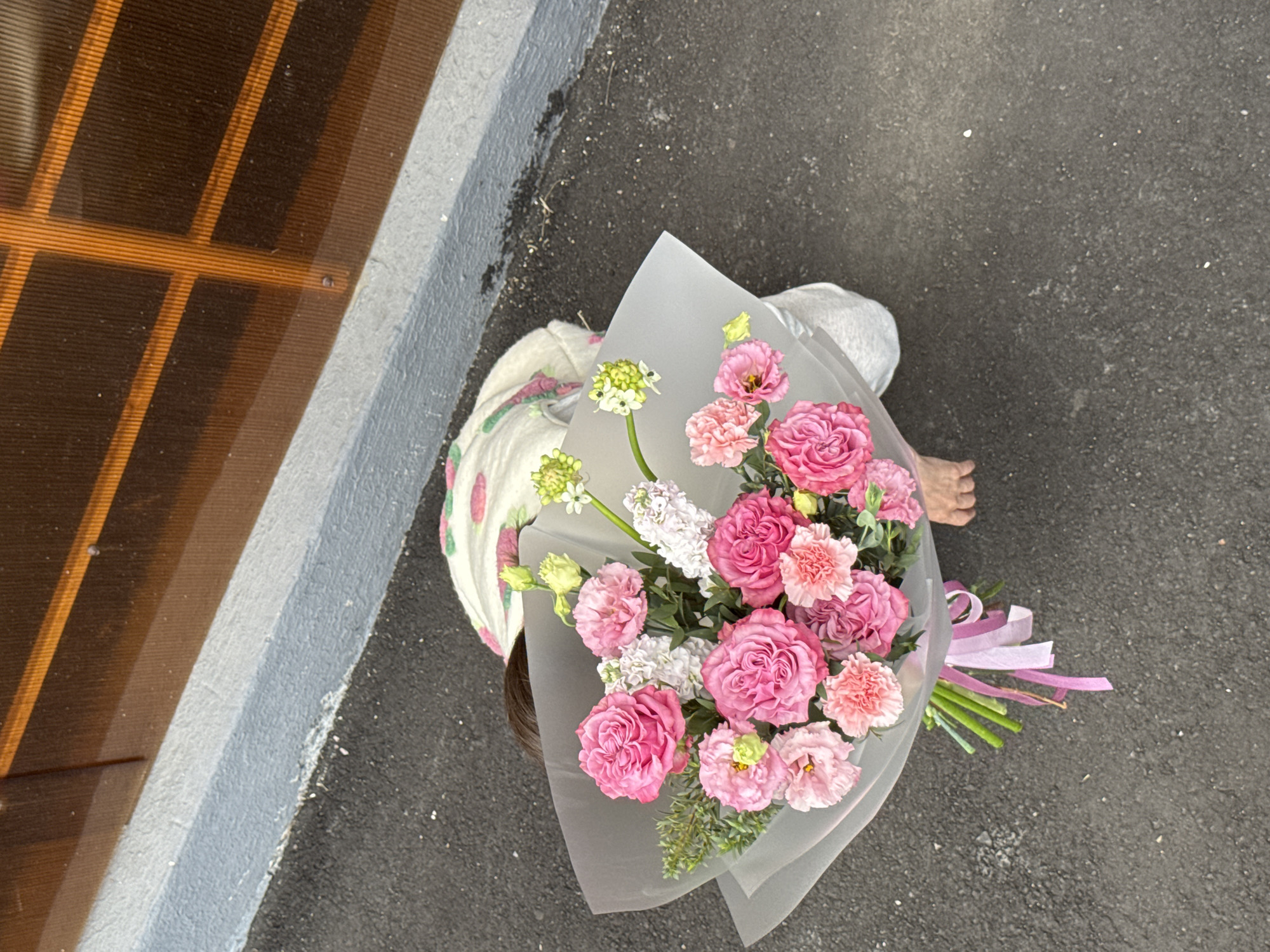 An overhead photograph shows a person holding a large, vibrant bouquet of flowers outdoors in Izhevsk, Russia. The person is partially visible, with only the top of their head and dark hair, a light-colored garment (possibly patterned), and one bare foot seen from above. They are standing on dark asphalt, which covers most of the ground. A grey concrete curb runs along the left edge of the frame, separating the asphalt from a dark, vertically striped surface, possibly a window or building panel.
The central focus is the substantial bouquet, which features a lush arrangement of pink and white flowers, interspersed with green foliage. Notable pink blooms include roses, carnations, and smaller, softer pink flowers (possibly lisianthus). White flowers, appearing in taller clusters, contribute to the bouquet's varied texture. The entire arrangement is wrapped in translucent, frosted-effect paper and tied at the stems with multiple trailing pink and white ribbons. The scene is brightly lit, suggesting it is daytime. No discernible text is visible in the image.