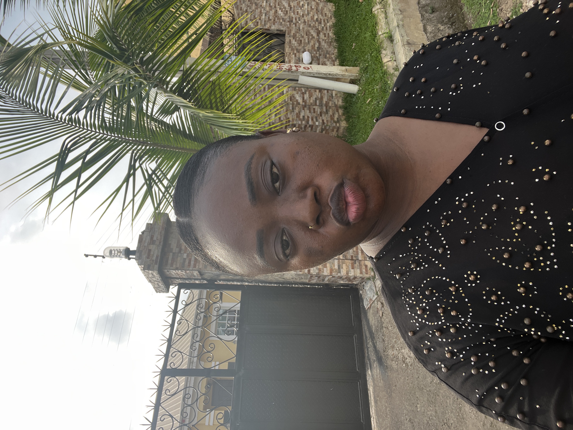 A person with dark skin and a nose ring is in the foreground, wearing a black top with decorative studs. Behind the person is a black metal gate with decorative patterns. A light-yellow building with white-framed windows is behind the gate. A brick pillar is on the left side of the gate, topped with a light fixture. A palm tree is visible to the left of the frame. The location appears to be a residential area in Abuja, Nigeria.
