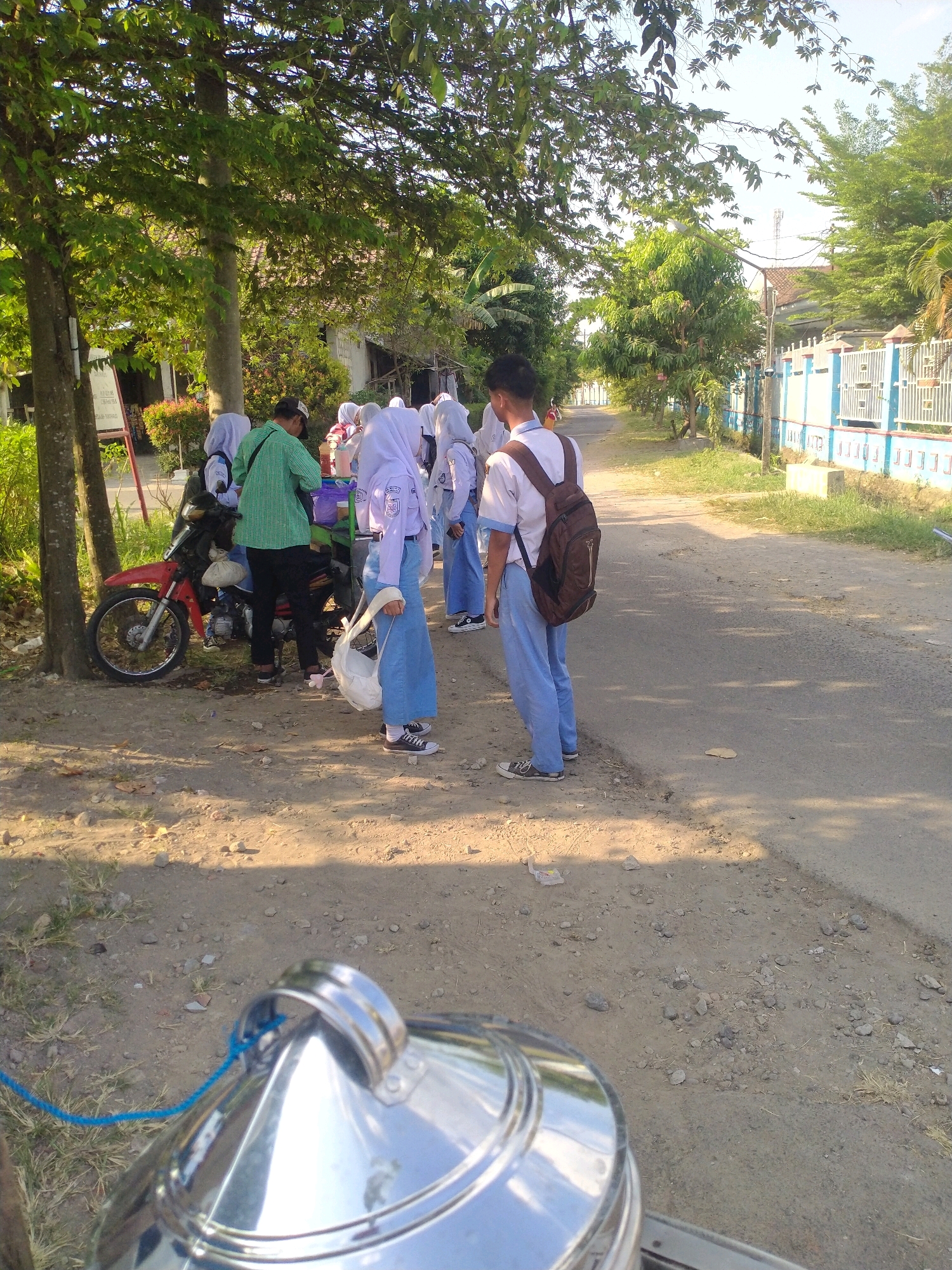 In Begajah, Indonesia, on a dirt and asphalt road bordered by trees and a white and blue fence, a group of students are gathered around a motorcycle. The students, wearing school uniforms consisting of white shirts, blue pants, and white hijabs, appear to be interacting with a person in a green shirt. One student in the foreground is carrying a white bag and wearing black sneakers. Another student, facing away from the camera, has a brown backpack and is also wearing blue pants and black sneakers. A red motorcycle is parked on the side of the road, with a silver object in the foreground, possibly part of a vehicle or container, secured with a blue rope. The scene is illuminated by sunlight, casting shadows on the ground.