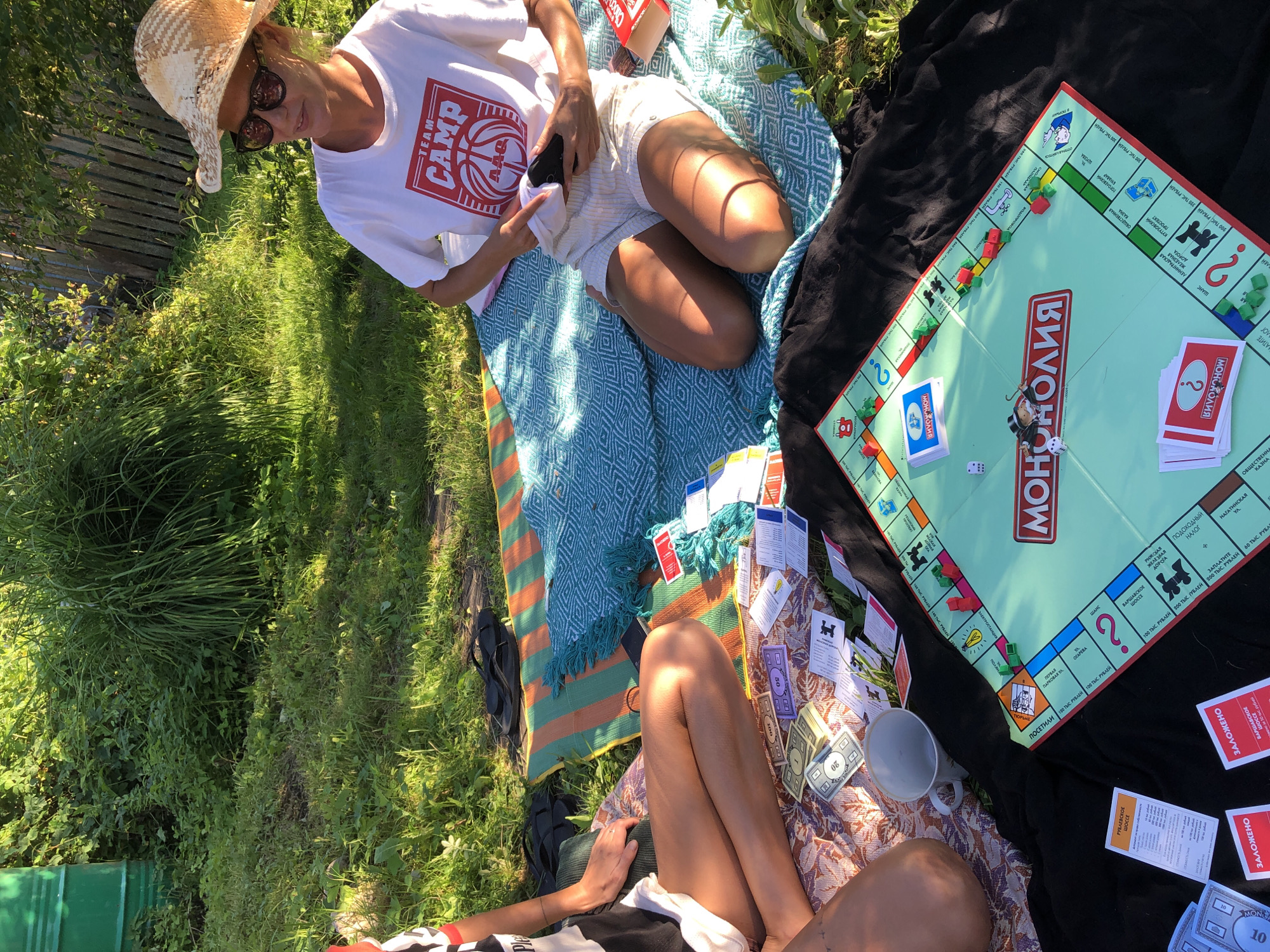 The image shows two people playing a game of Monopoly outdoors on a grassy area. A woman wearing a straw hat and sunglasses is seated on a blue and green patterned blanket, holding a phone in her hand. She is wearing a white t-shirt with "TEAM CAMP" printed on it and striped shorts. To her left, another person, partially visible, is also seated on the blanket. The Monopoly board is set up on a black cloth spread on the ground, with various cards and fake money scattered around it. The background consists of lush green foliage and a wooden fence, suggesting a garden or park setting. The lighting indicates it is a sunny day.