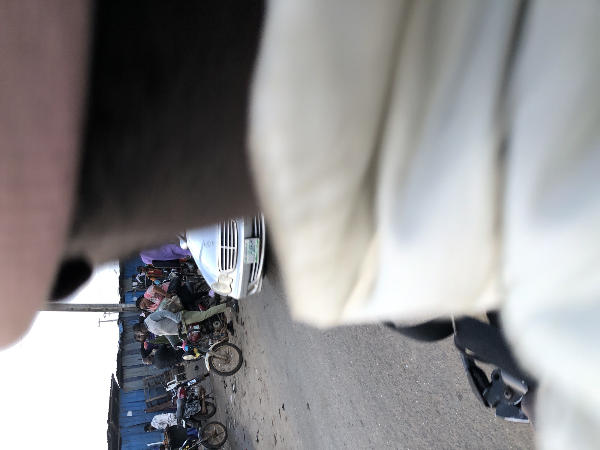 The image depicts a busy street scene in Ikeja, Nigeria, likely a market or transportation hub. In the foreground, a blurry view of a person wearing a light-colored jacket is visible, suggesting the photo is taken from their perspective, possibly on a motorcycle. Several motorcycles are parked or in motion, with some people interacting with them. One motorcycle in the mid-ground has a person in a grey shirt and green trousers seated on it. Further back, a silver car is seen driving on the road. The background features blue corrugated metal structures and a utility pole. The ground is asphalt, littered with some debris. The overall impression is of a vibrant, somewhat chaotic urban environment.
