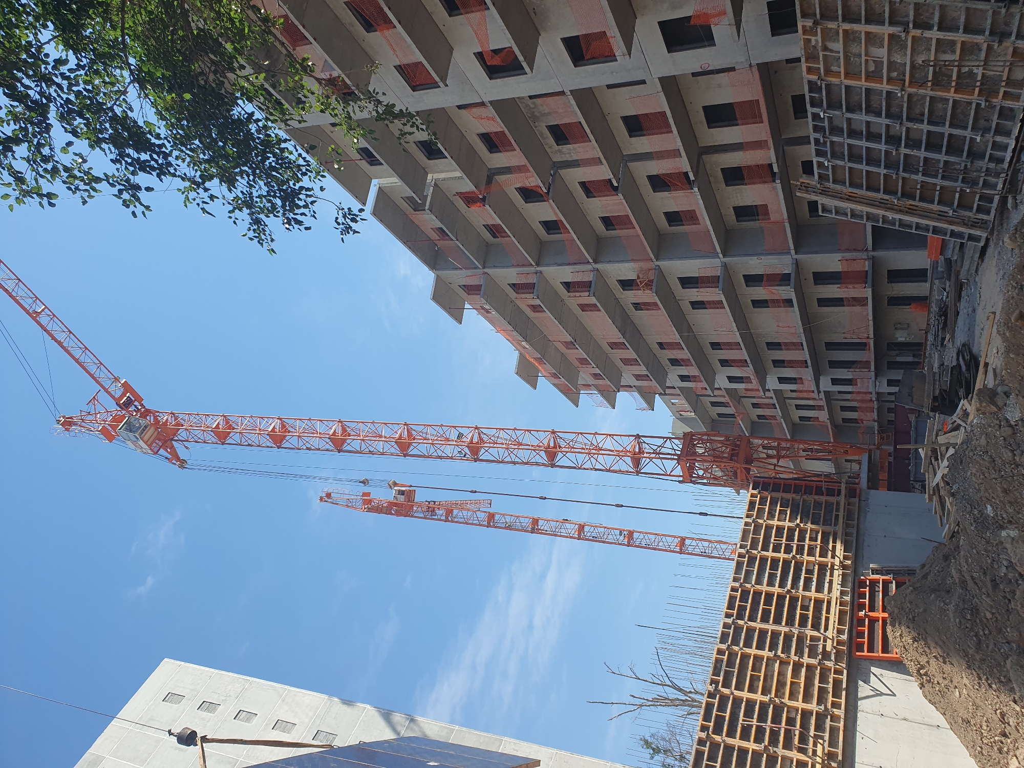 The image depicts a construction site under a clear blue sky, likely in Tashkent, Uzbekistan, given the architectural style of the building and the presence of multiple tower cranes. The primary focus is on a multi-story concrete building in various stages of construction, characterized by exposed concrete floors, balconies with orange safety netting, and dark window openings. Two large orange tower cranes dominate the skyline, one significantly taller than the other. In the foreground, there is a concrete foundation with wooden formwork, piles of dirt, and construction materials. Another partially constructed building or wall made of concrete is visible to the left. The scene suggests an active construction project in progress.